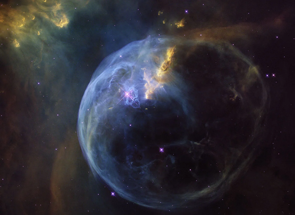 The Bubble Nebula, also known as NGC 7635, is an emission nebula located 8,000 light years away in the Cassiopeia constellation. The bubble is created by stellar wind pushing out from a massive hot central star, visible here just left of the center of the image, a purple star partly obscured by blue gas clouds. The nebula is seven light years across—about one-and-a-half times the distance from our sun to its nearest stellar neighbor, Alpha Centauri. This stunning image was observed by the NASA/ESA Hubble Space Telescope to celebrate its 26th year in space.