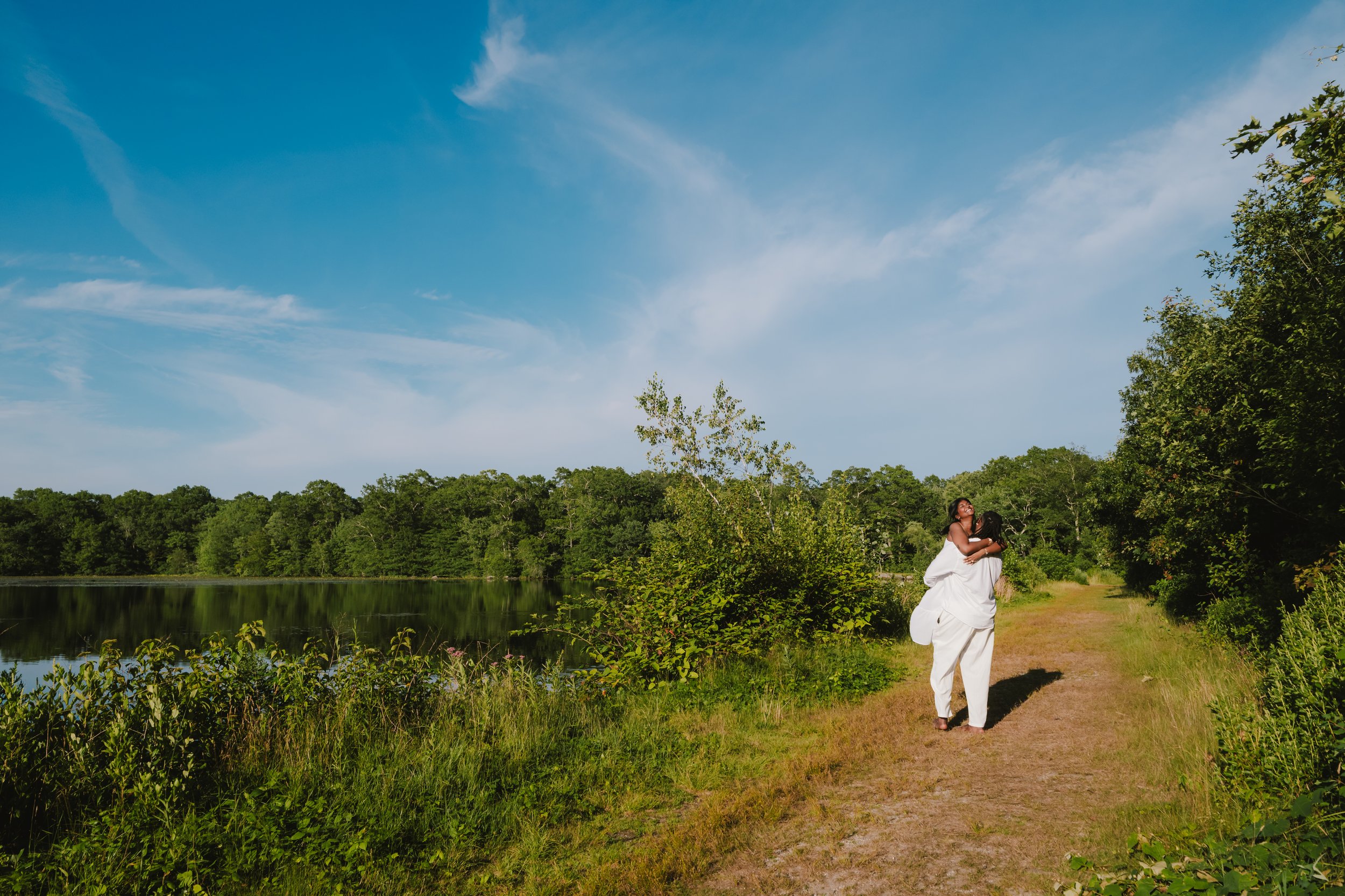 Two women in white outfits embracing on a dirt path near a lake, surrounded by green trees and a blue sky with some clouds.