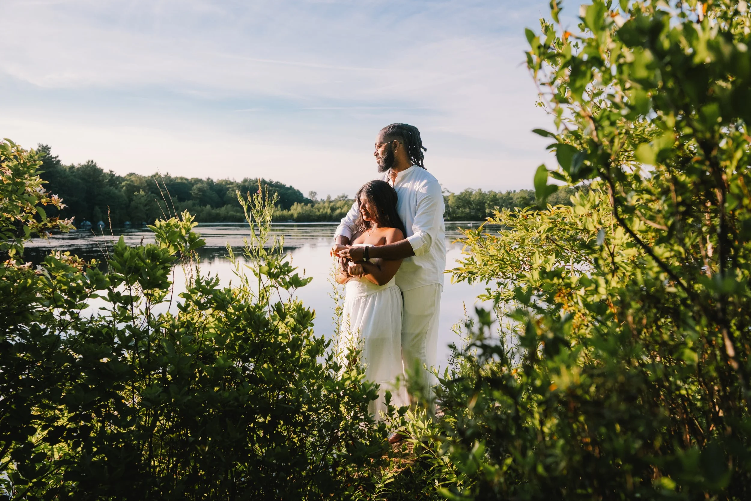 A couple stands by a lake surrounded by lush greenery in the late afternoon sunlight, with the woman holding a smartphone.