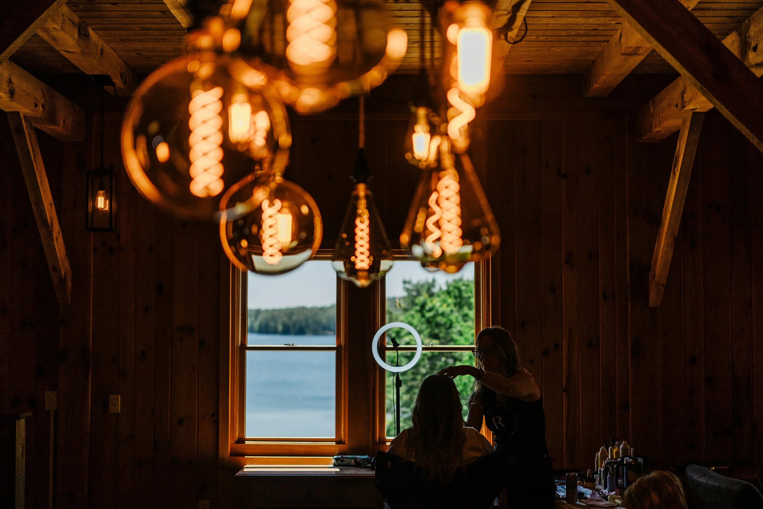 A bride is silhouetted in front of a window while she gets her hair and makeup done. Decorative light bulbs are seen on the ceiling in the near foreground during a Maine wedding.