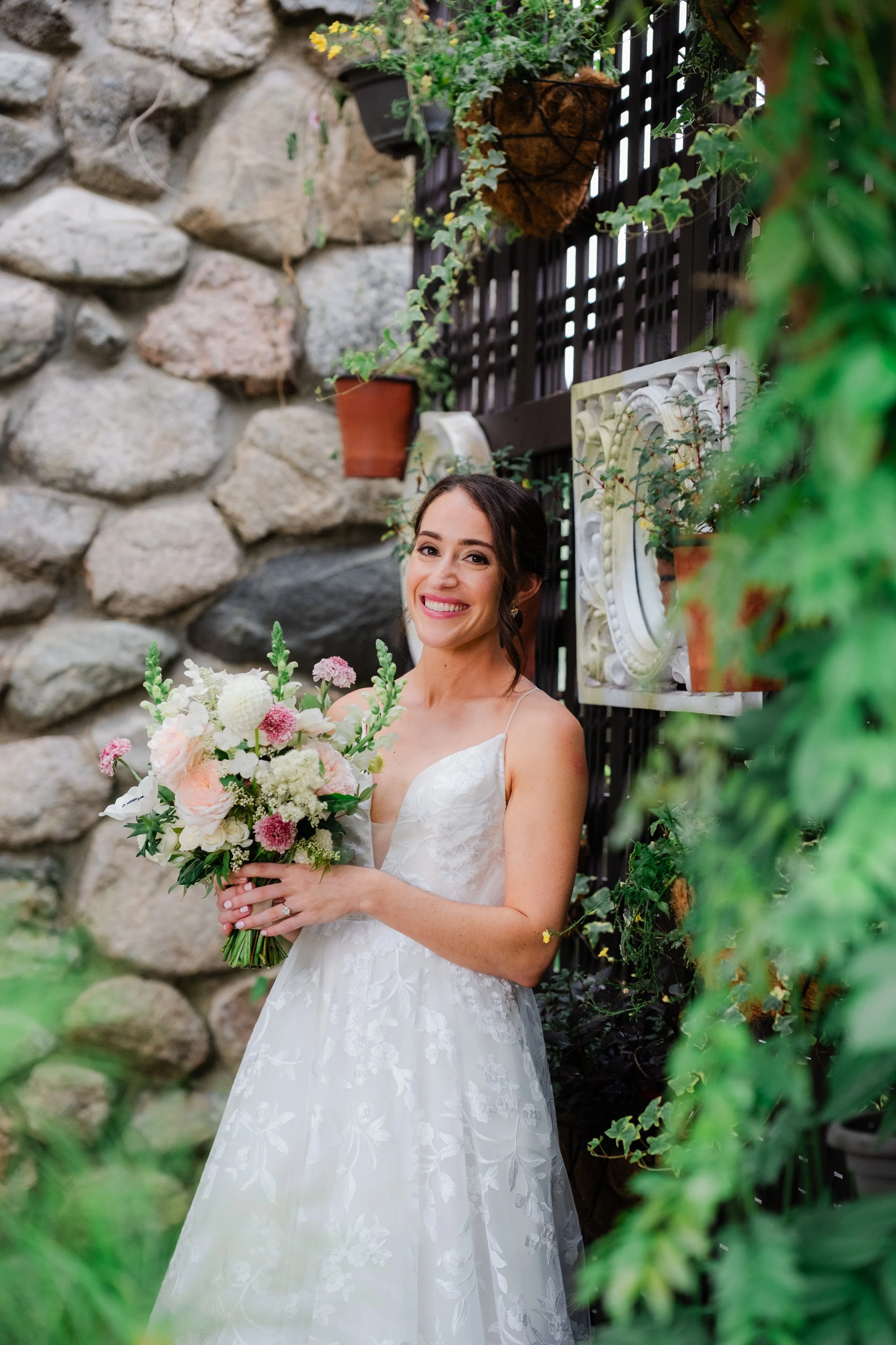 A smiling bride in a white wedding dress holding a bouquet of flowers, standing in a garden with stone walls and hanging pots of greenery and flowers.