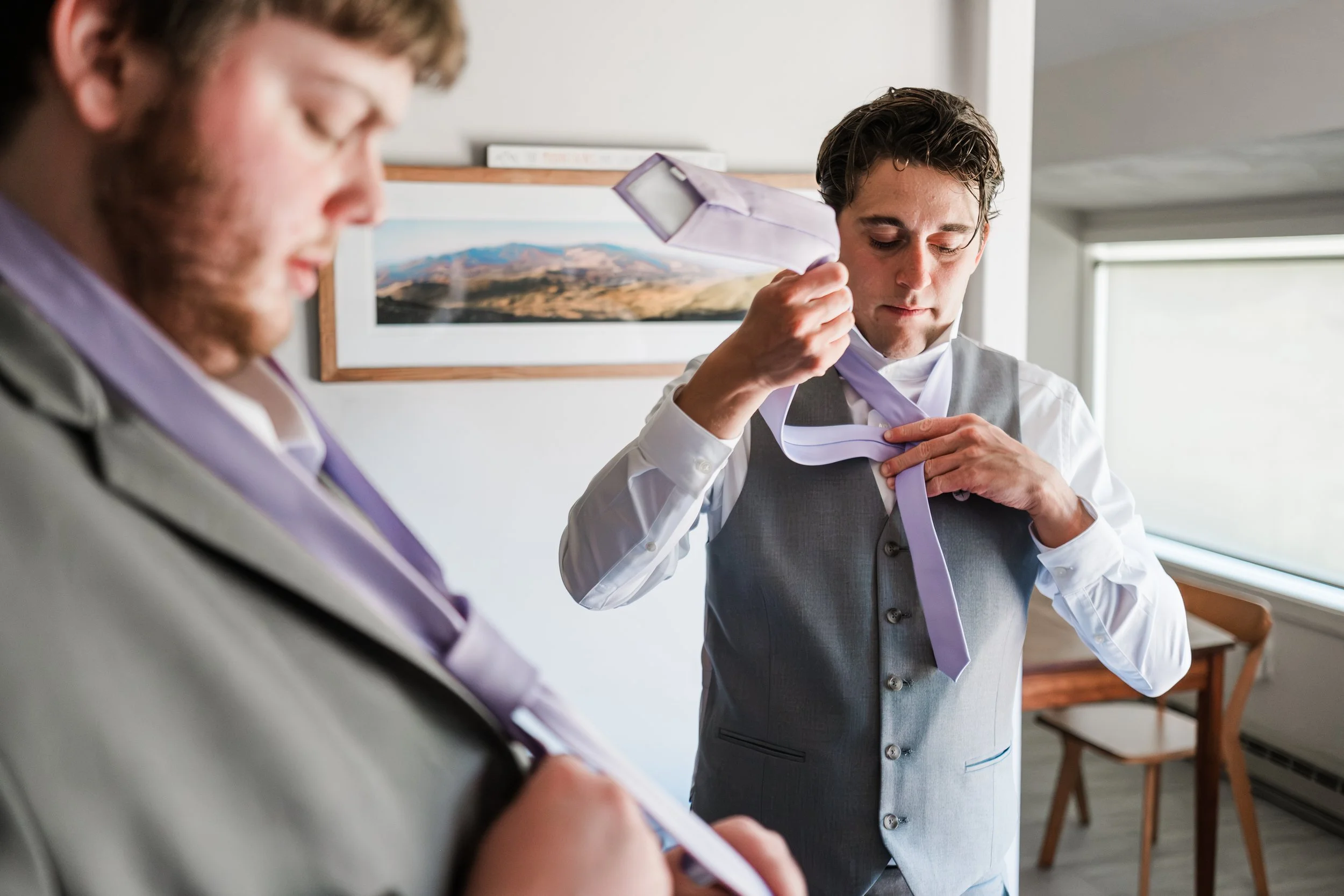 Two men in formal attire, one adjusting a lavender tie, the other looking at a paper, in a bright room with a mountain picture on the wall.
