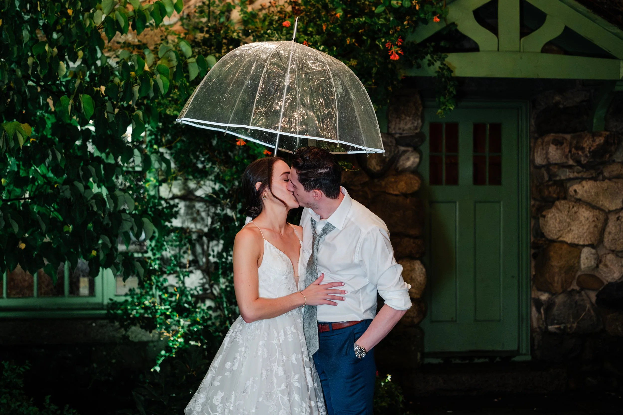 A couple sharing a kiss under a clear umbrella in the rain, with greenery and a stone building in the background.