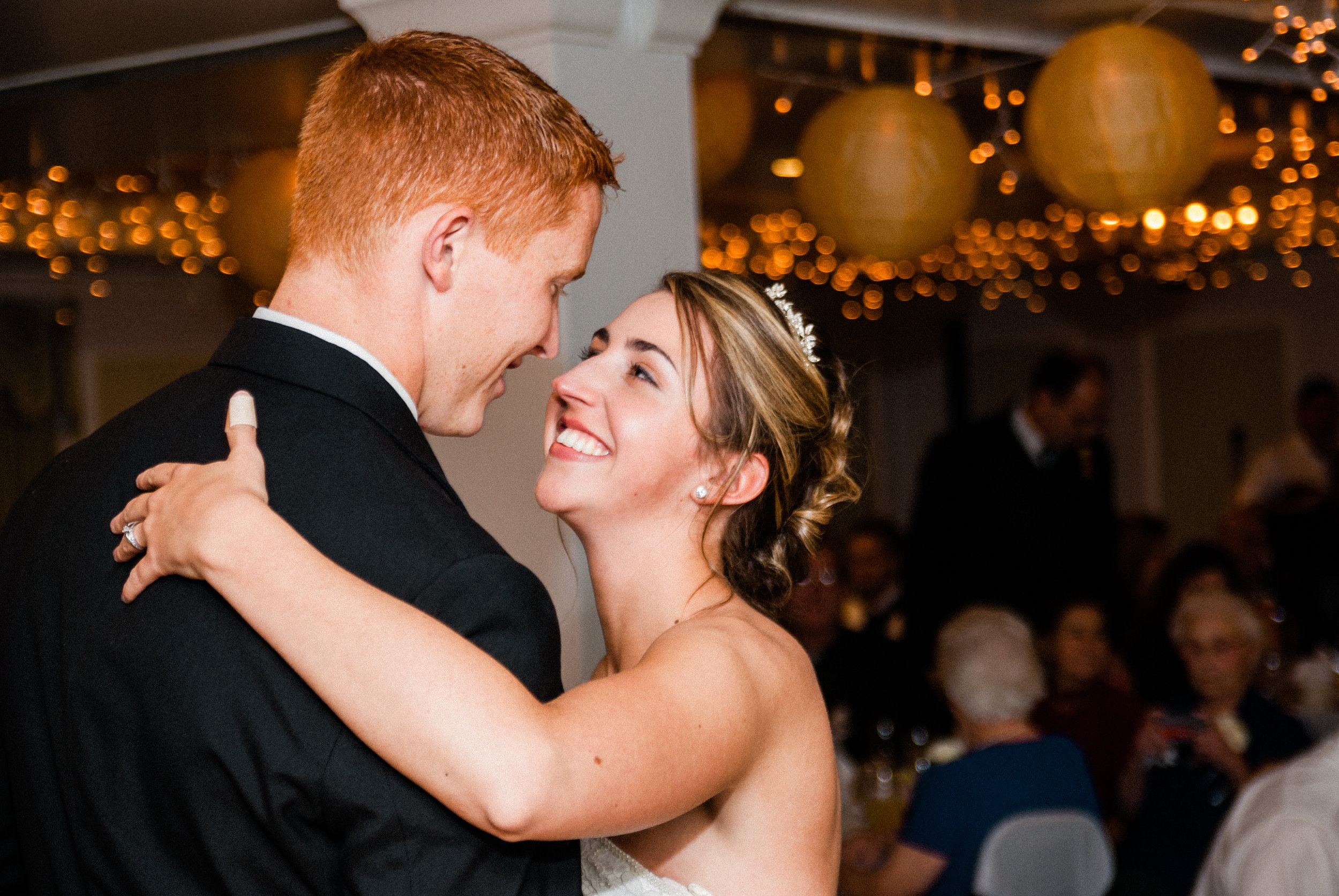 A newlywed couple dances closely together at their wedding reception, smiling and gazing into each other's eyes. The bride has curly hair styled in an updo with a small tiara, and the groom has short red hair. In the background, guests sit at tables 