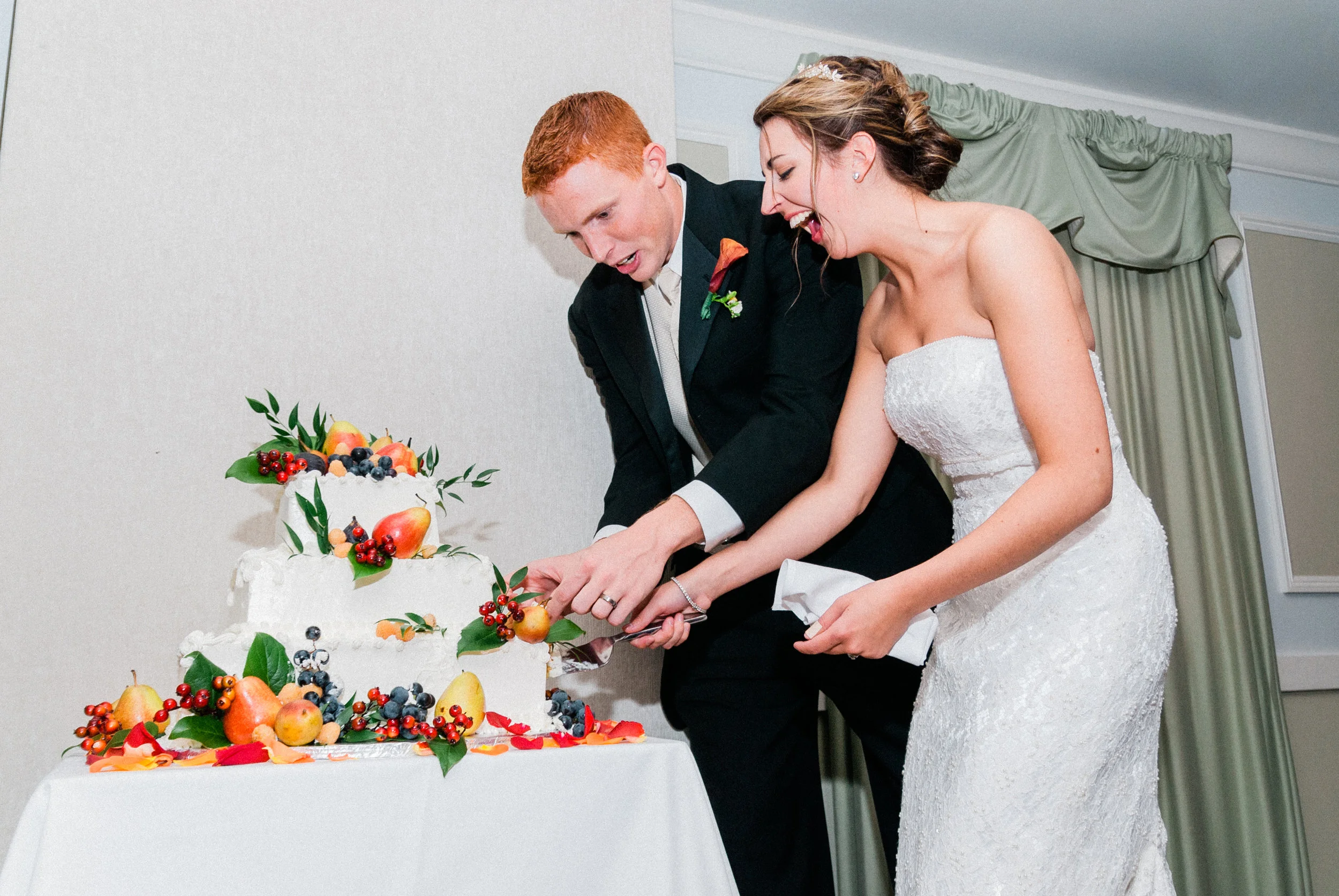 A couple in wedding attire cutting a wedding cake decorated with fruit and leaves, smiling and celebrating.