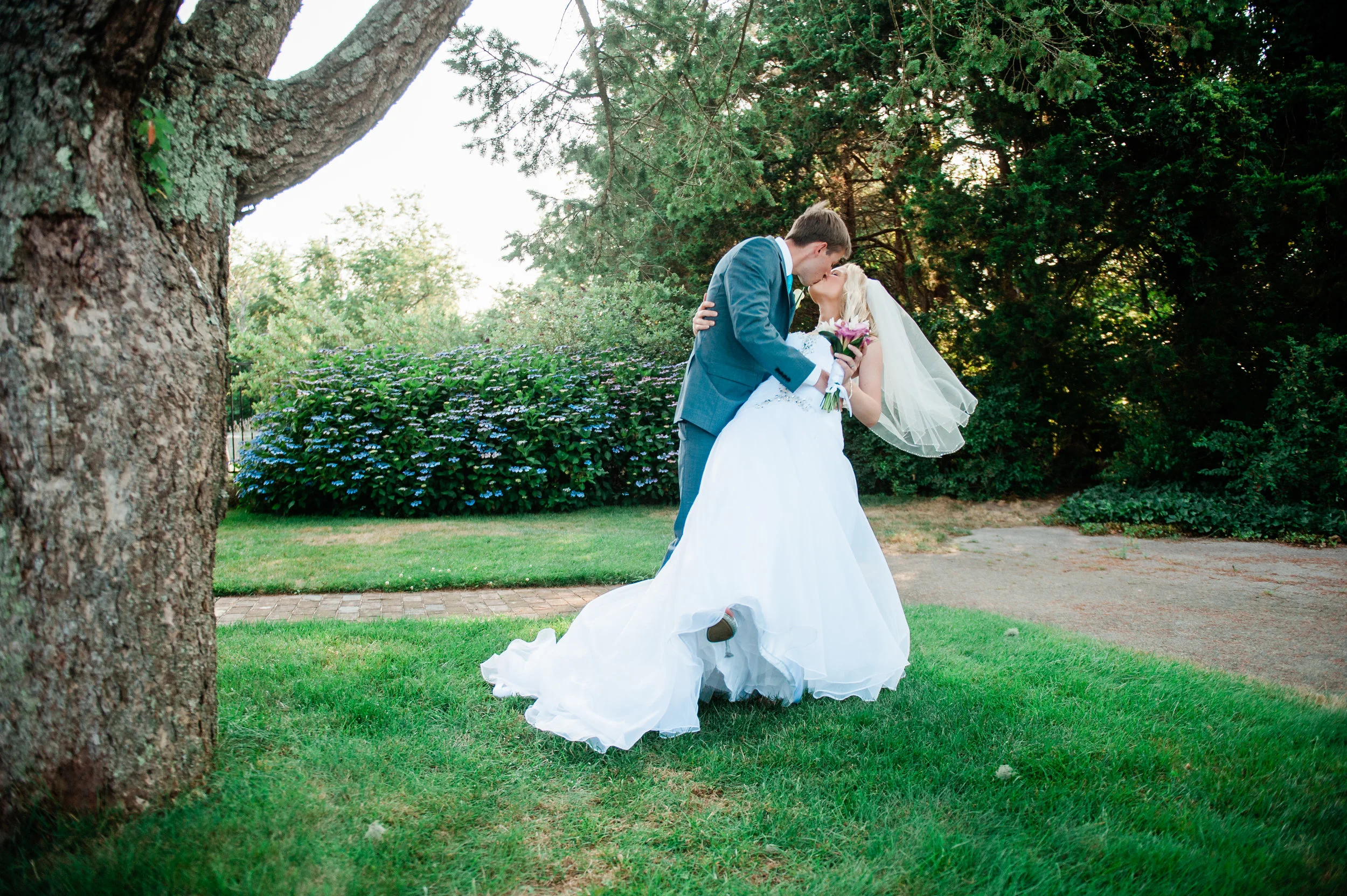 A bride and groom sharing a kiss outdoors on a grassy area during their wedding, with trees and bushes in the background.