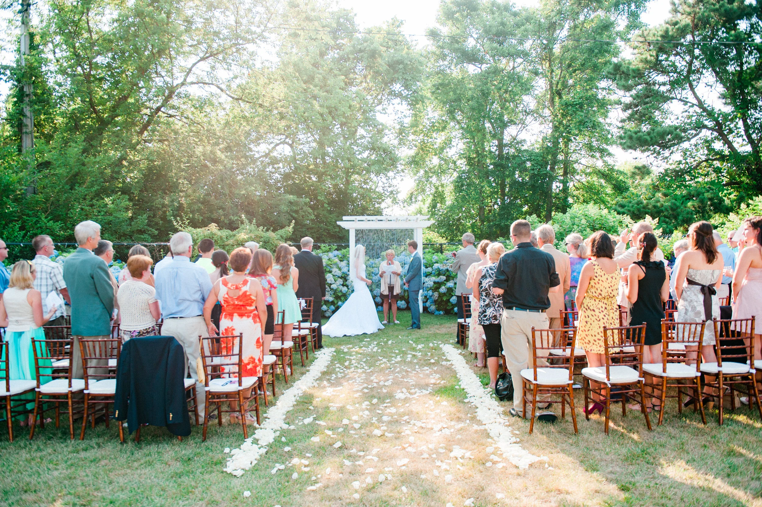 An outdoor wedding ceremony with people gathered around a bride and groom, with a white arch and floral decorations surrounded by tall trees.