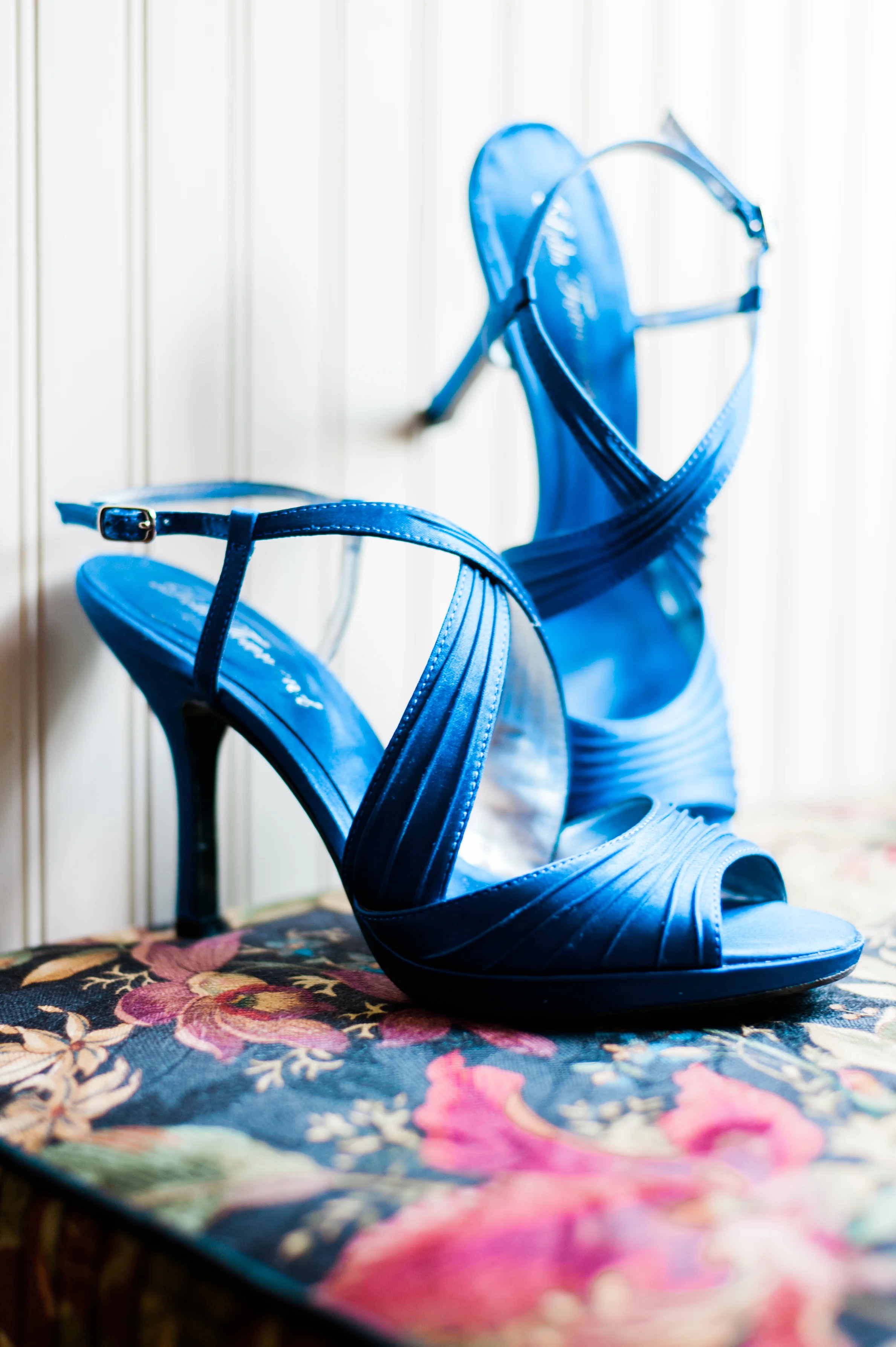 A pair of shiny blue high-heeled sandals with straps, placed on a floral patterned cushion.