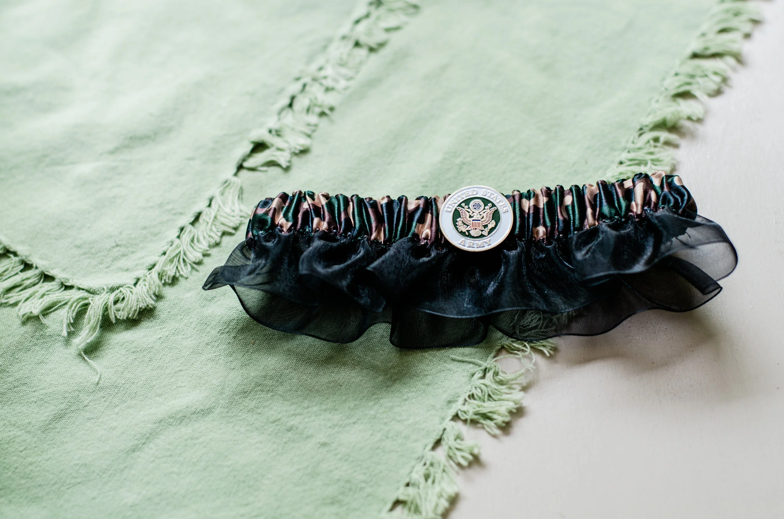 A black leather bracelet with a colorful striped elastic band and a United States Army emblem, decorated with black sheer fabric ruffles, resting on a light green fringed fabric.