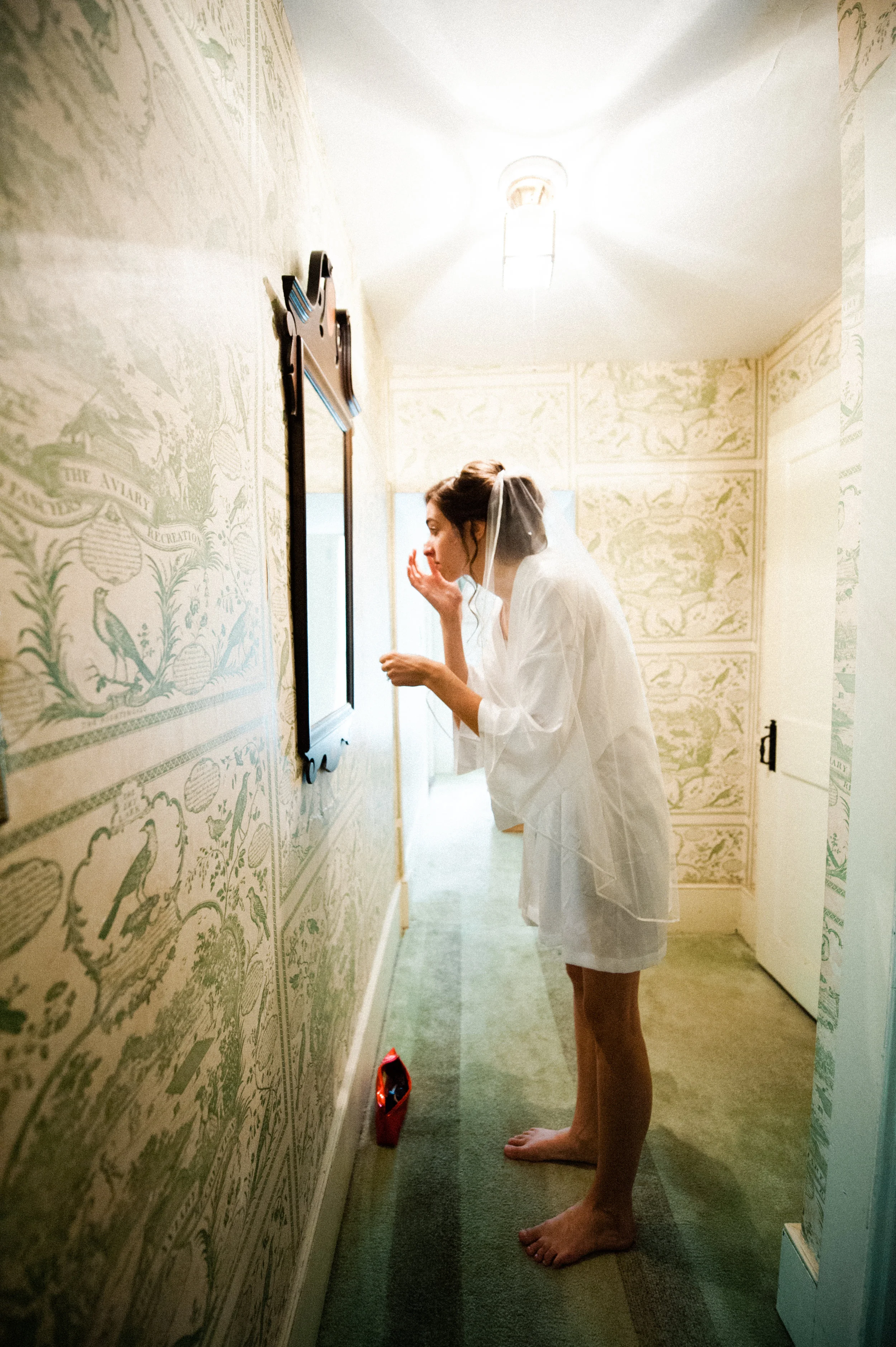 A woman in a white robe with a veil is looking at herself in a mirror on a wall inside a room with patterned wallpaper, applying makeup or skincare.