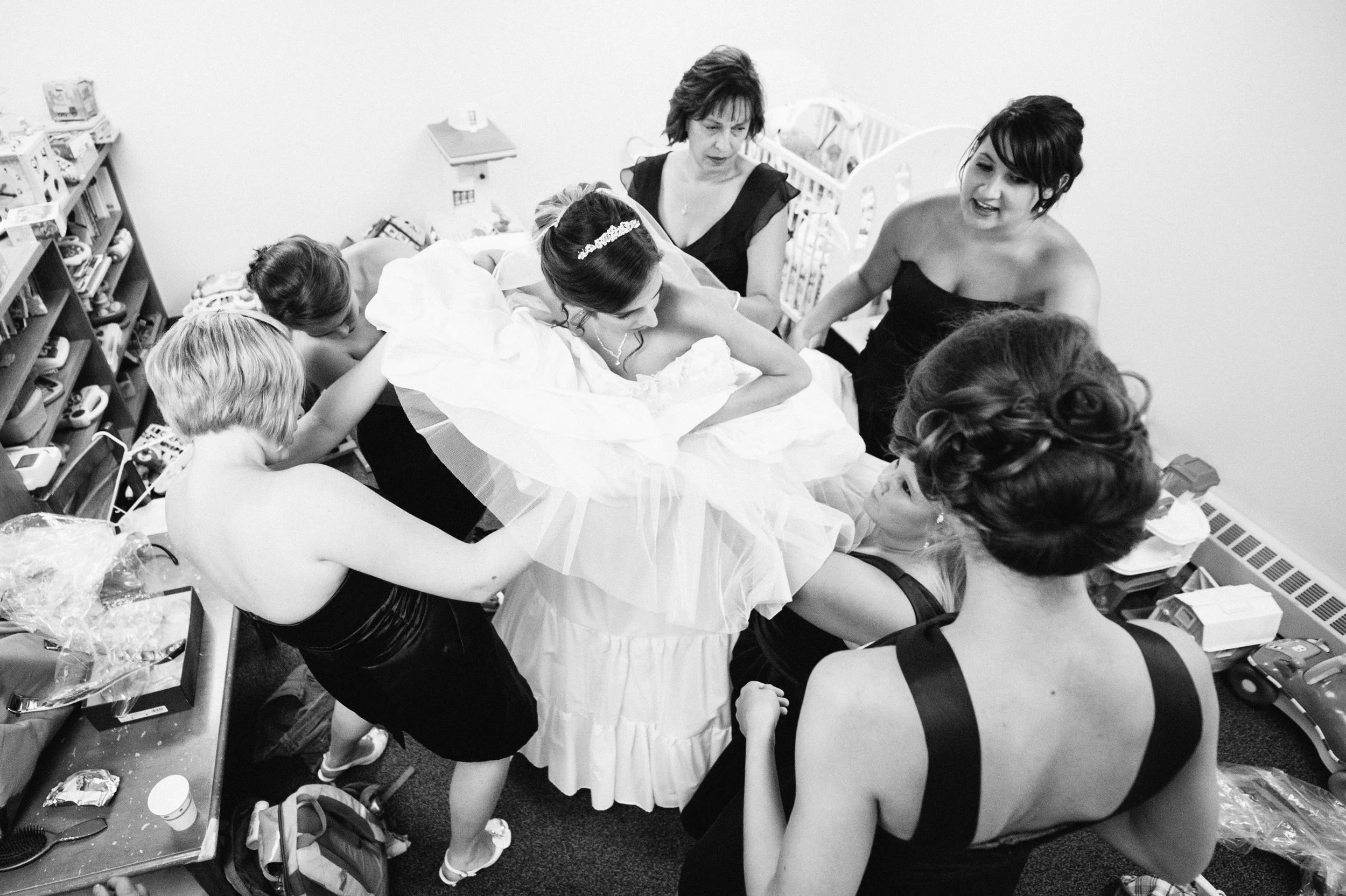 A bride is surrounded by women helping her prepare, in a cluttered room with shelves of books and toys, as she leans on a pillow with a veil on her head.