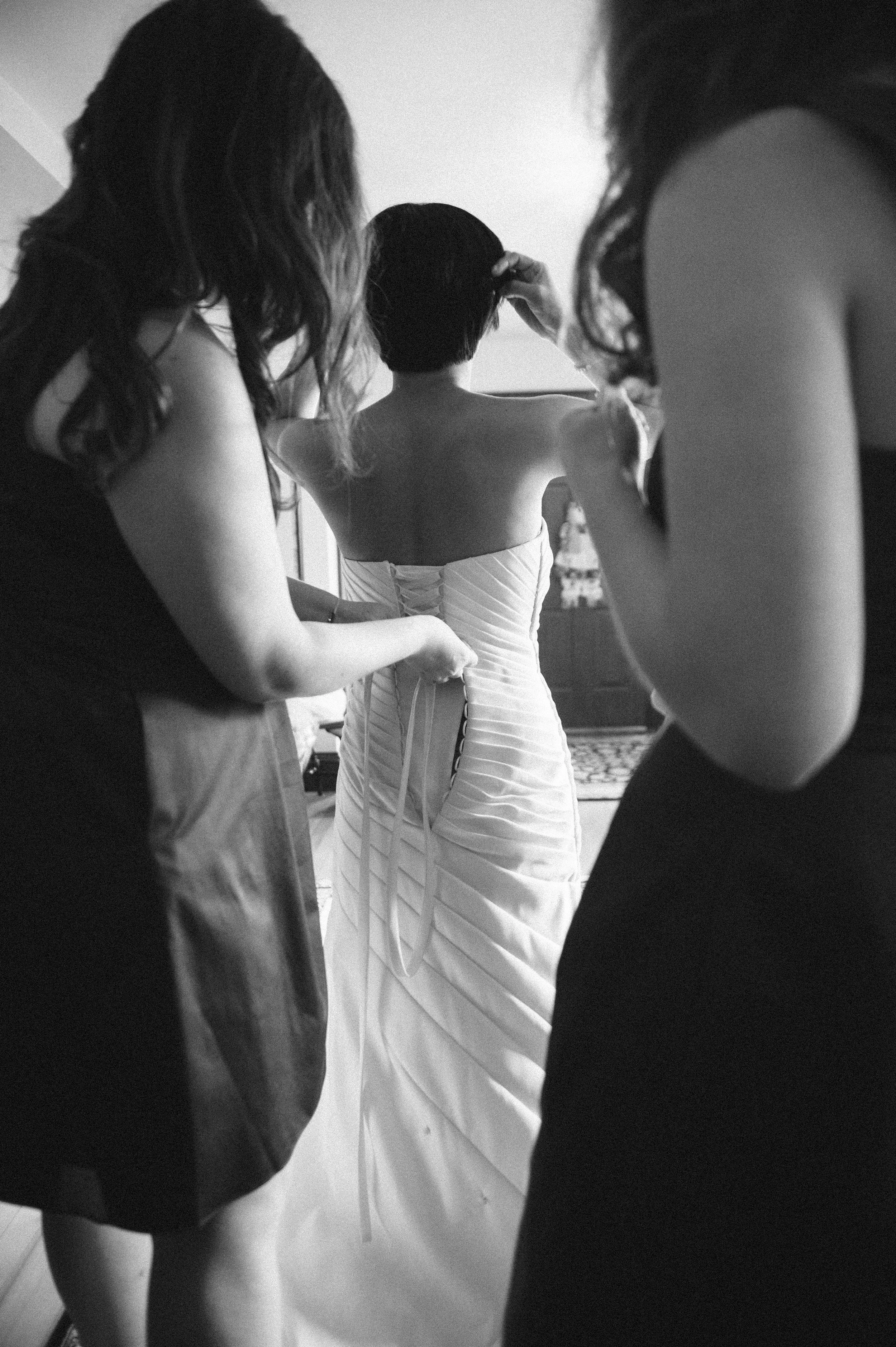 Three women helping a bride get ready, with the bride putting on a wedding gown in a room.