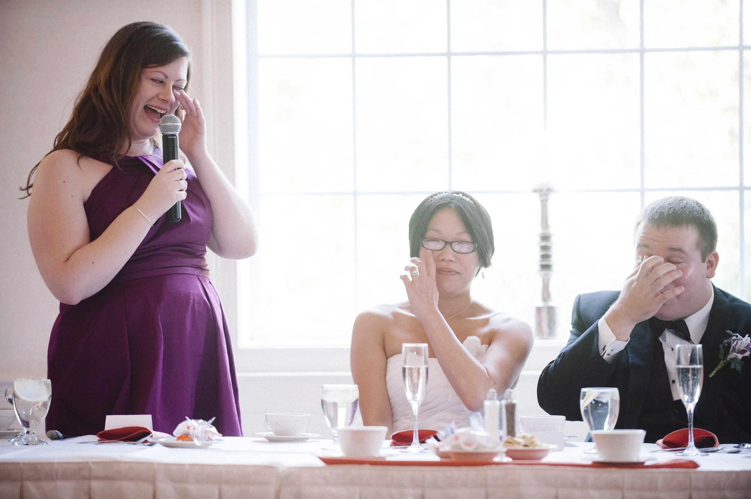 A woman in a purple dress giving a speech at a wedding reception, with a bride and groom seated at the table. The bride is adjusting her glasses, and the groom is holding his face with his hand. The table has glasses of champagne, cups, and plates.