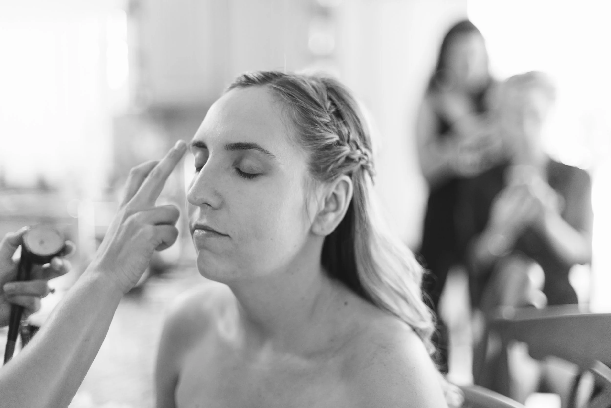 Woman with closed eyes having makeup applied to her face by a makeup artist in a bright room.