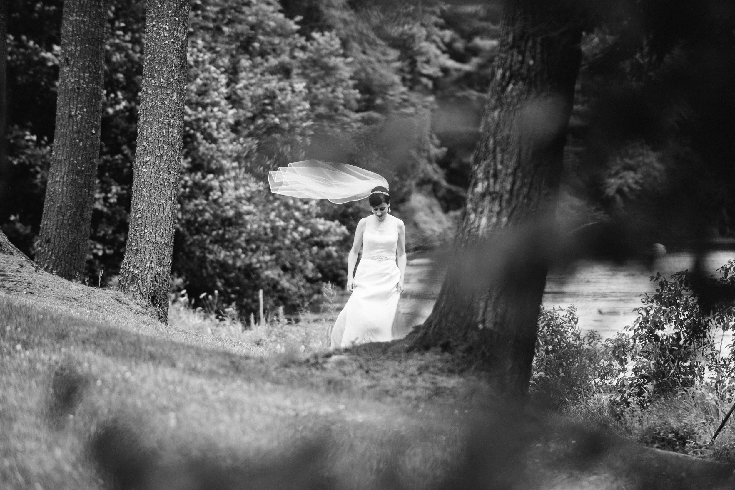 A woman in a sleeveless white dress walking through a wooded area with trees and a body of water nearby, with her veil flowing behind her.