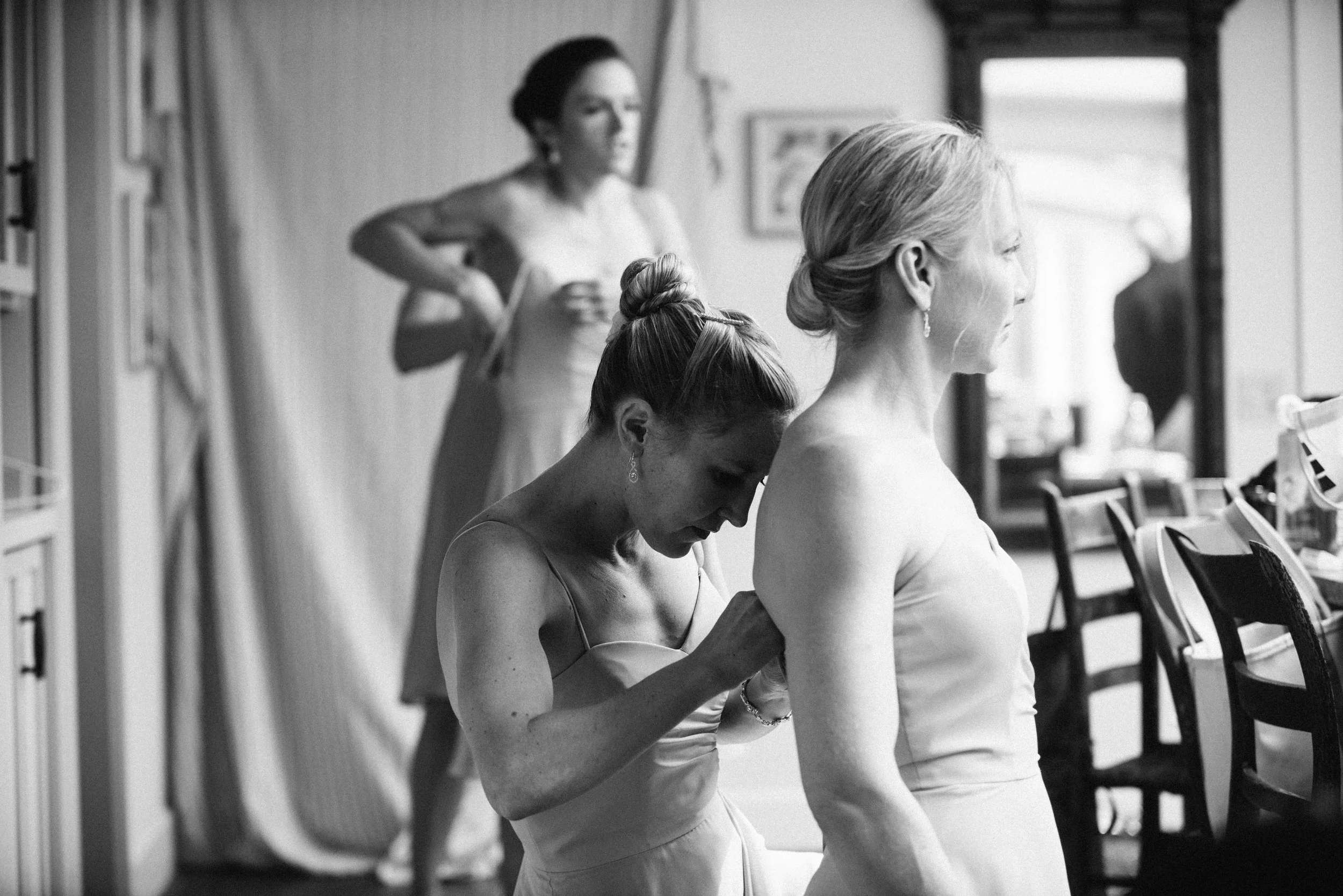 Four women in a room, with one adjusting and helping another woman prepare, possibly for a wedding, as they wear formal dresses and seem to be getting ready.