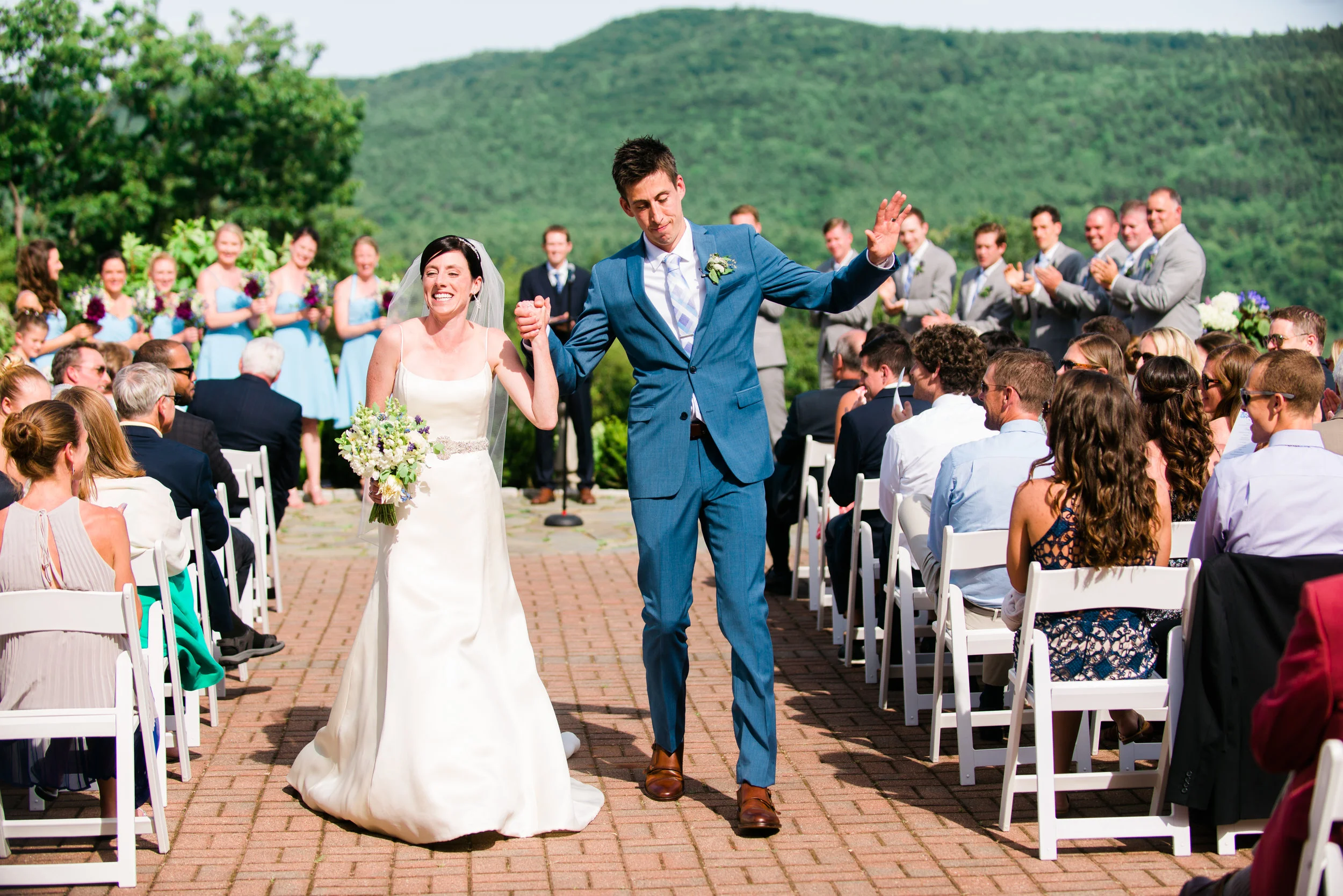 A wedding ceremony outdoors with a bride and groom walking down the aisle, holding hands, smiling. The bride is in a white wedding dress holding a bouquet, the groom is in a blue suit. Guests are seated on white chairs, clapping and smiling, with a g
