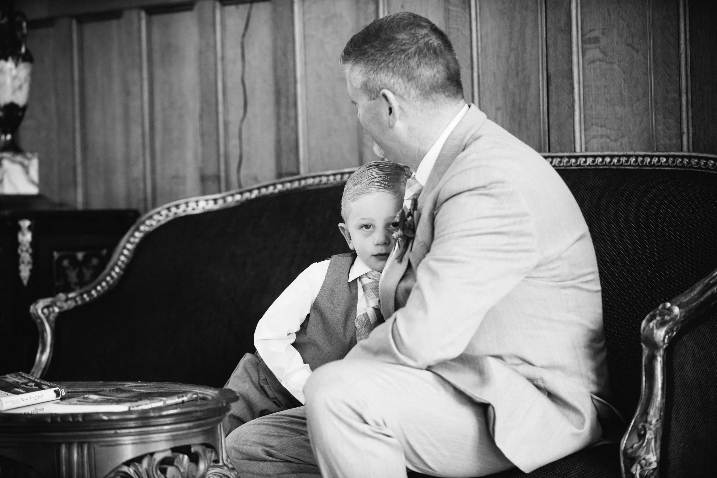 A man and a young boy sitting together on a vintage couch in a wood-paneled room. The boy is leaning against the man, looking directly at the camera, while the man is looking at the boy. Both are dressed in formal attire, with the boy in a vest and t