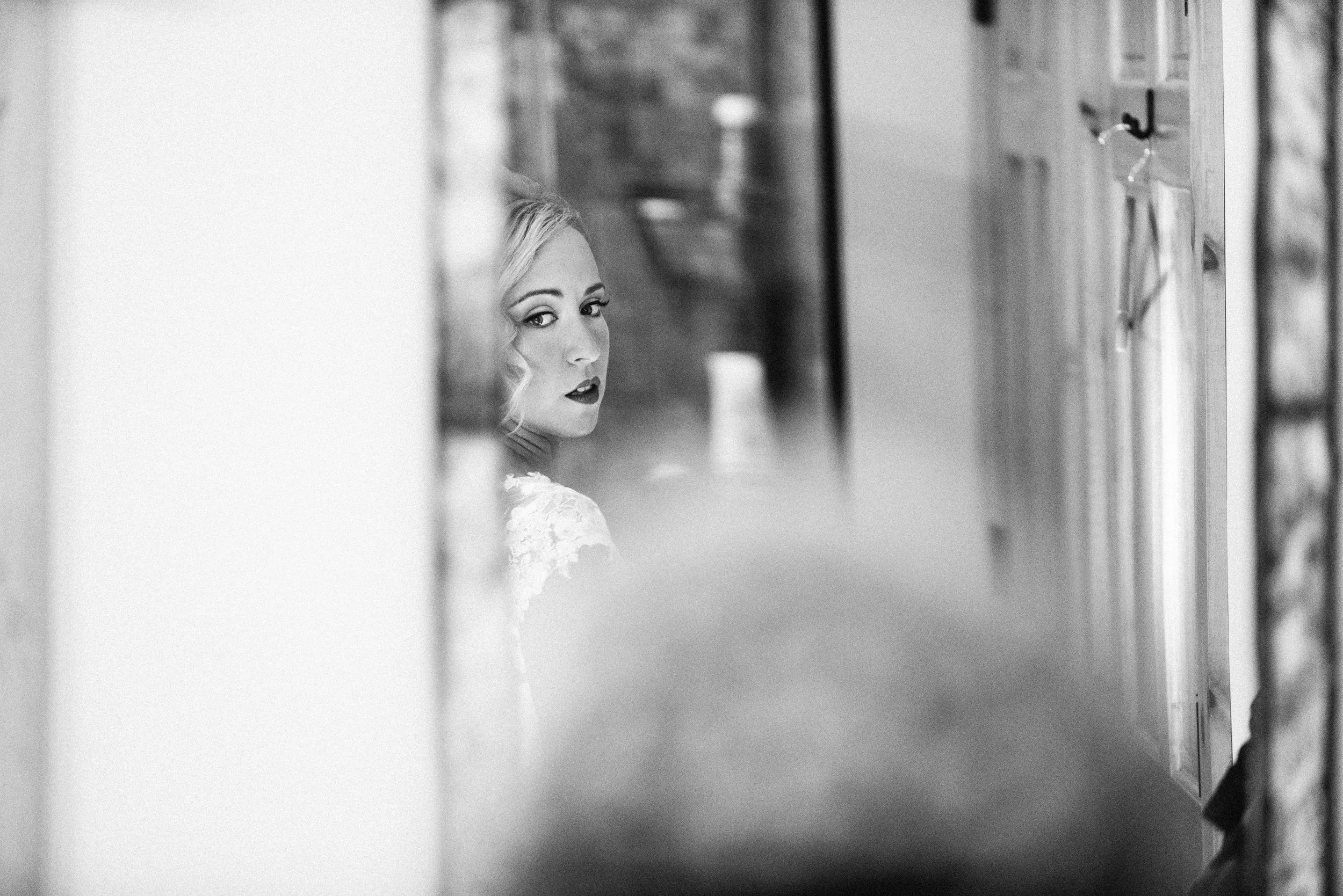 A black-and-white photo of a woman with styled hair and makeup, looking towards the camera through a narrow gap.