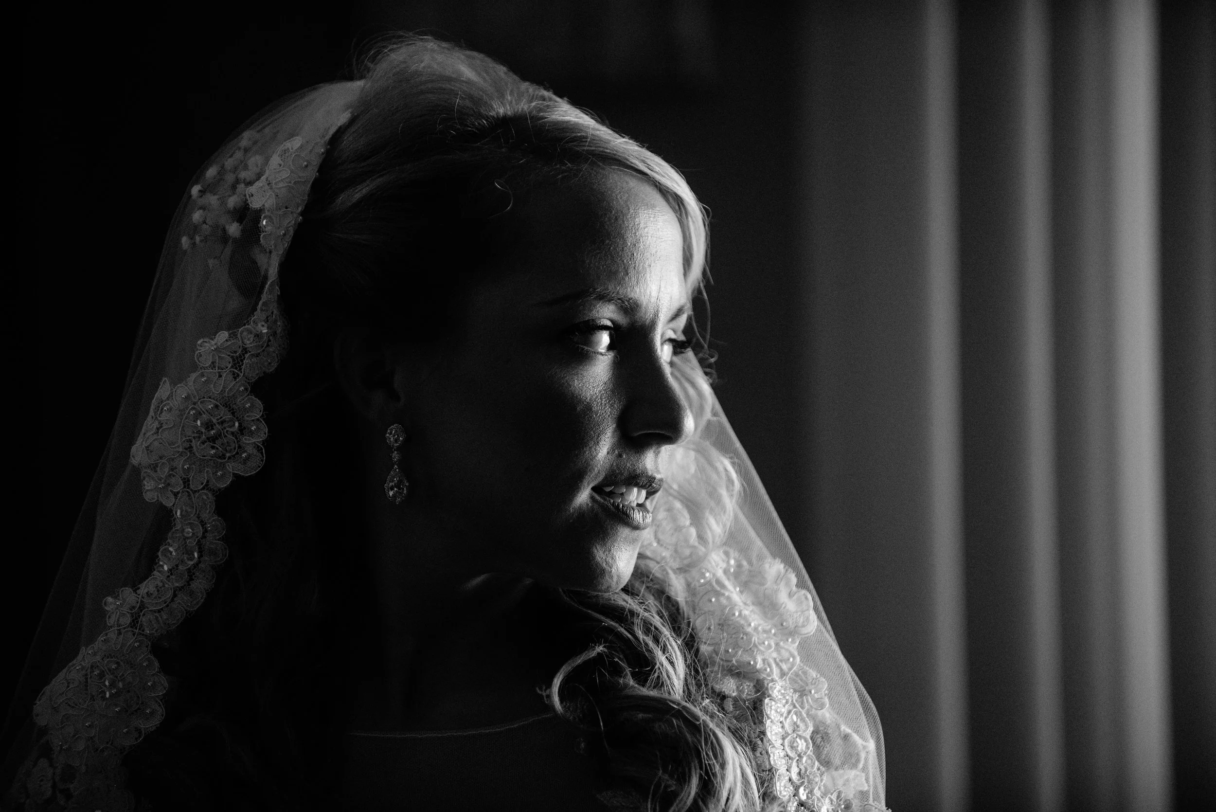 A bride looking out a window, wearing a lace veil and earrings, with soft lighting highlighting her face.