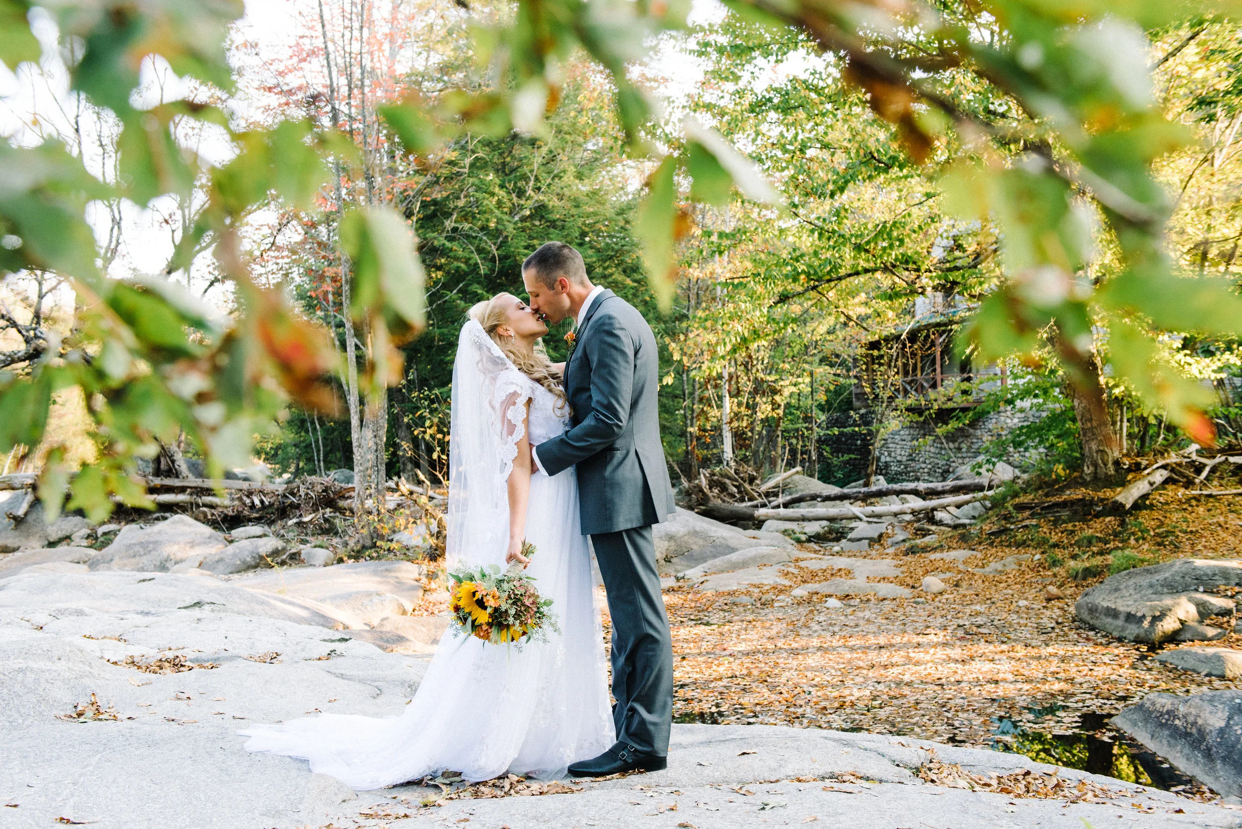 A bride and groom embrace on a rocky outdoor trail during autumn, surrounded by colorful fall foliage, with a rustic cabin in the background.