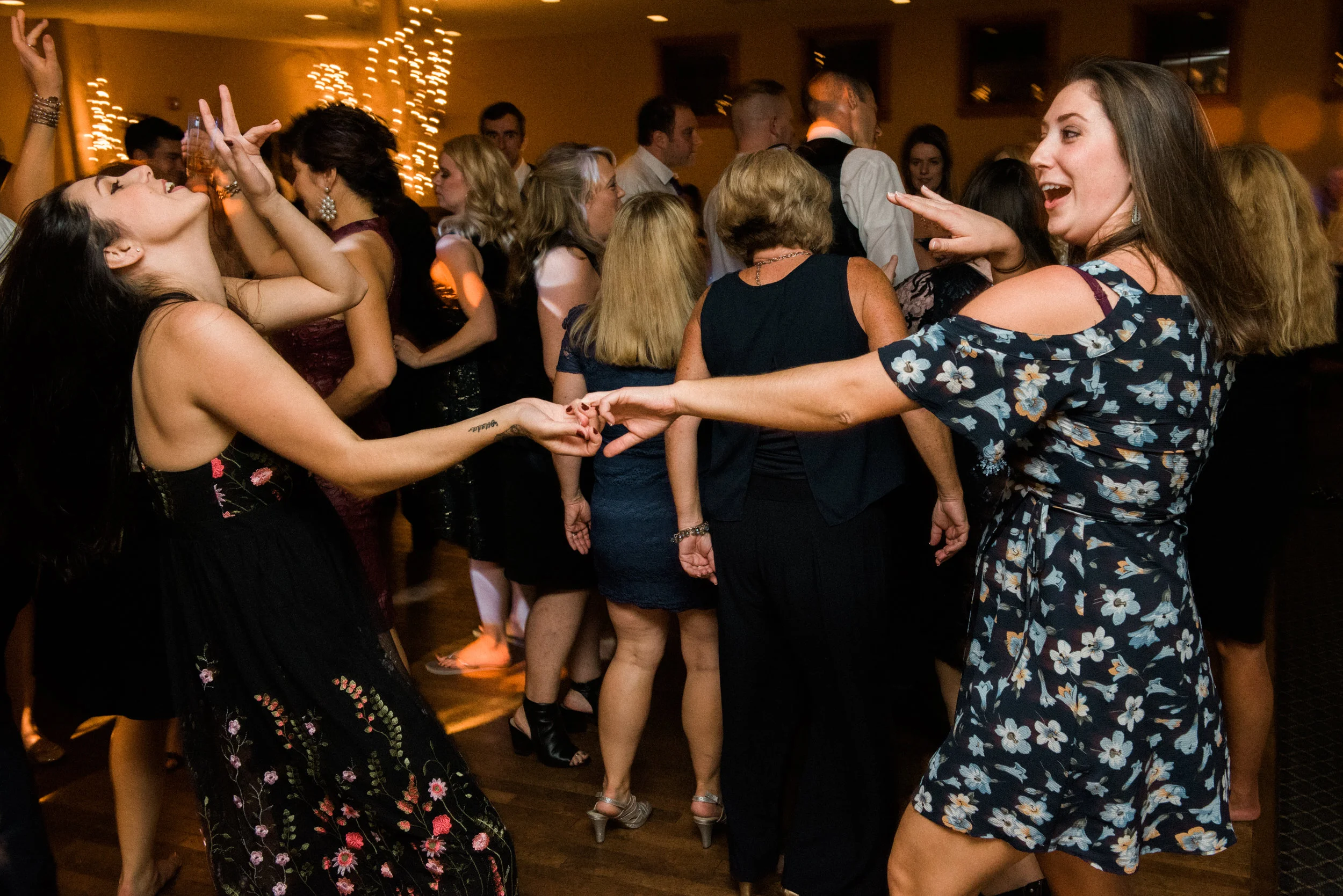 Two women dancing and holding hands at a party with other people dancing in the background.