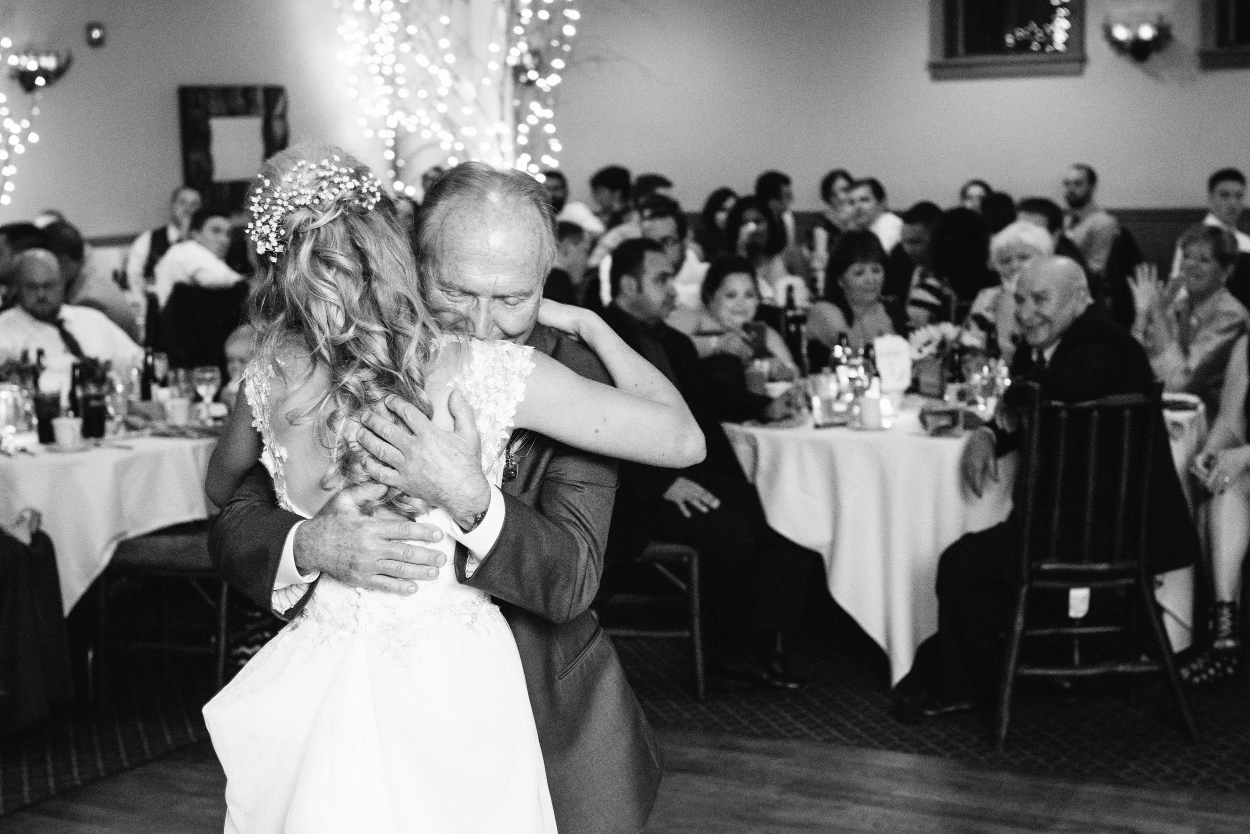 A bride and an older man, likely her father, dancing and hugging at a wedding reception, with guests seated at tables in the background.