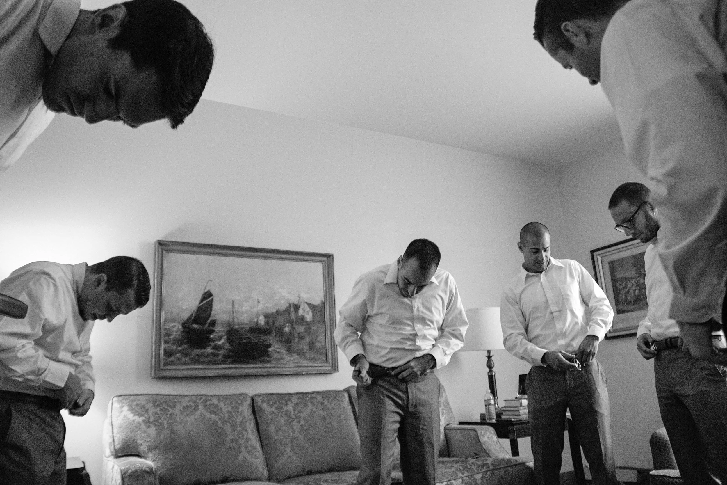 Group of five men in white shirts and dress pants getting ready or adjusting their clothing in a living room, with paintings, a lamp, and a couch in the background.