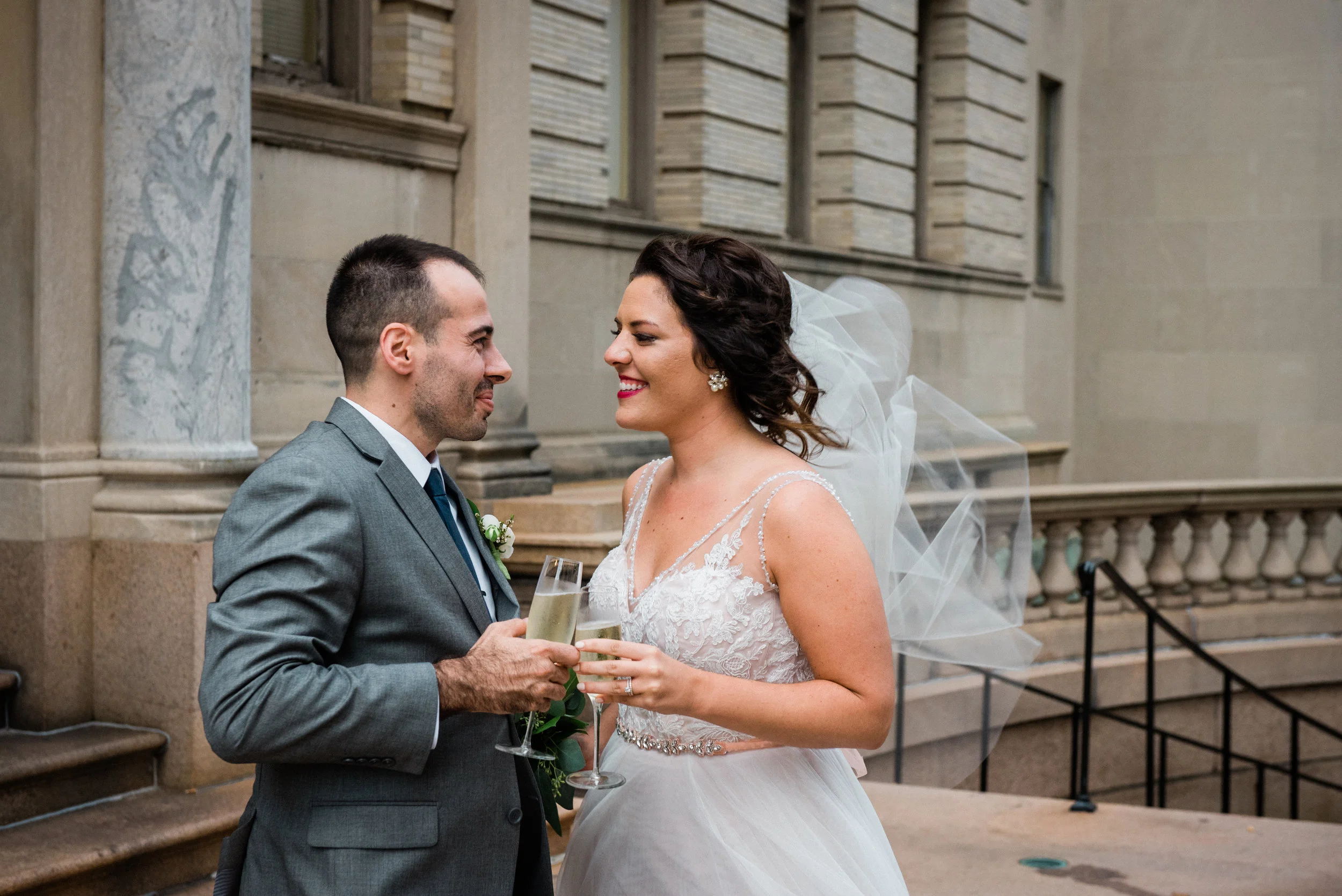 Bride and groom in wedding attire holding champagne glasses and smiling at each other outside a stone building.