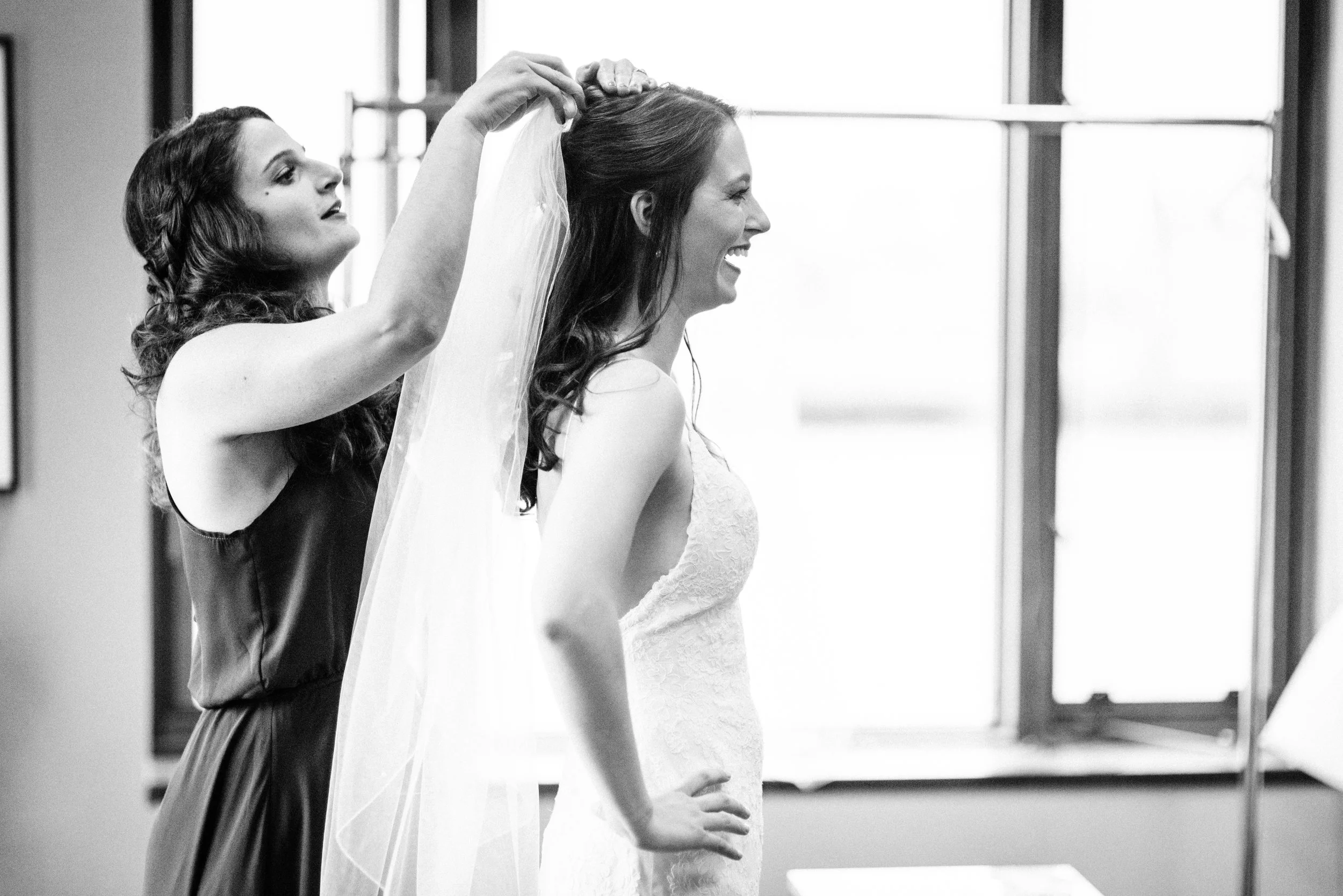 A woman in a wedding dress smiling as she is helped with her veil by another woman.