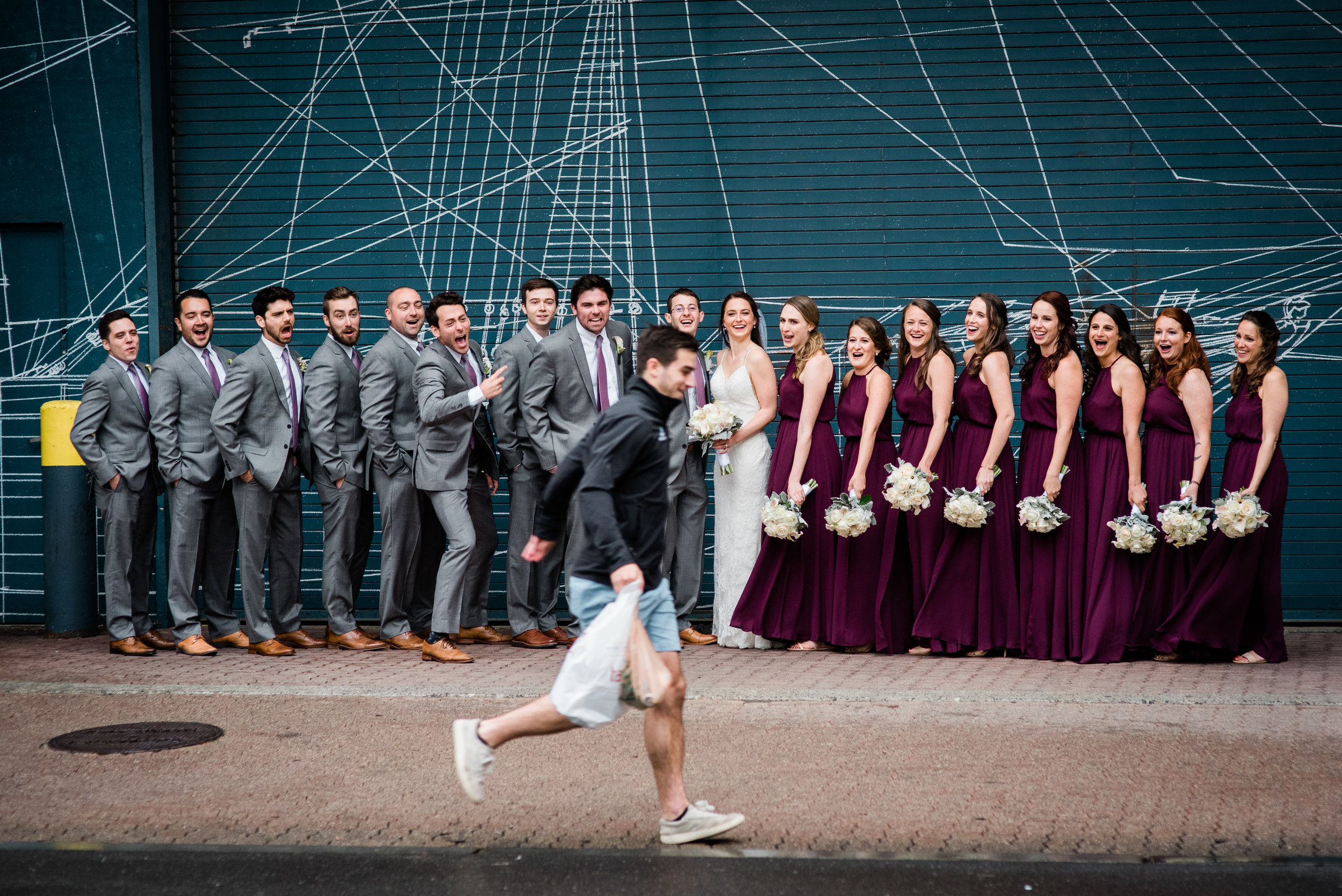 A group of bridesmaids and groomsmen posing for a photo in front of a blue wall with white geometric designs. The bride and groom are in the center, smiling and holding bouquets of flowers. A man is running in front of the group, wearing casual cloth