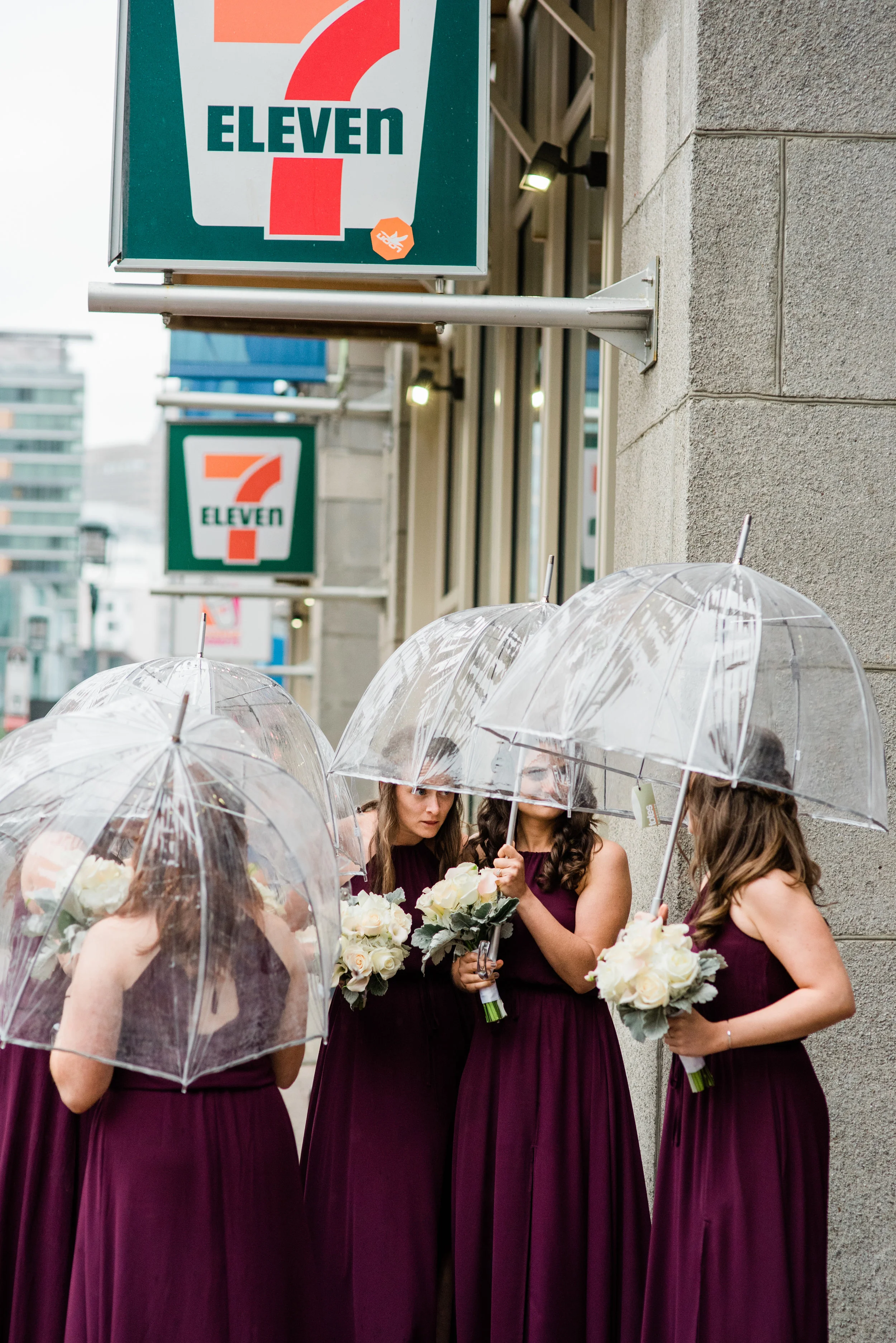 Four women in matching purple dresses holding bouquets of white flowers, standing under clear umbrellas outside a 7-Eleven store on a rainy day.