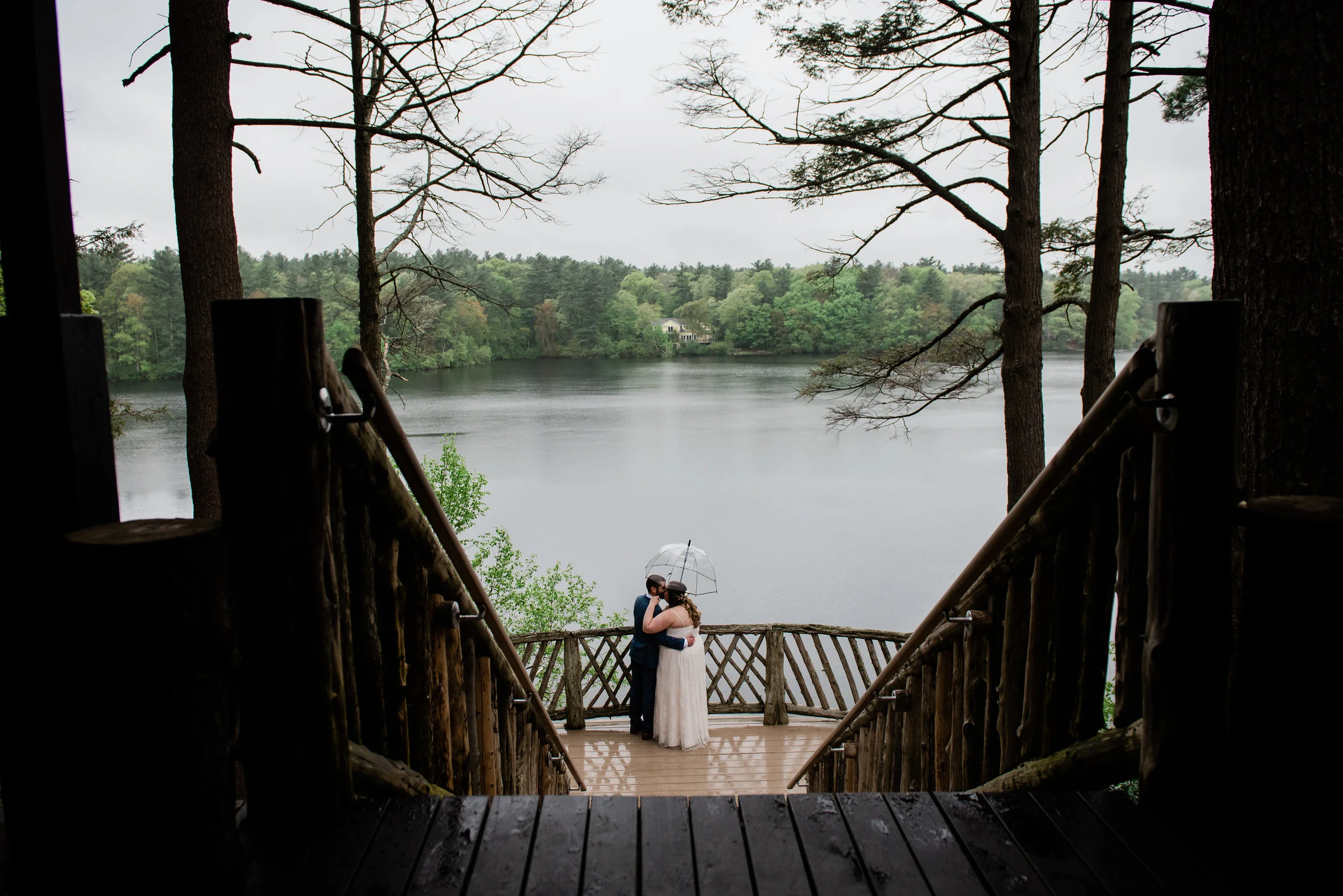 A couple in wedding attire standing on a wooden dock under an umbrella, embracing by a lake with tall trees and a cloudy sky in the background.