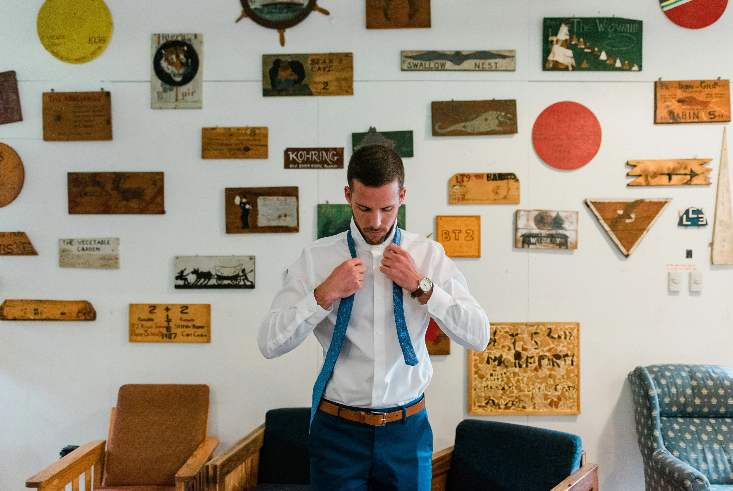 A man wearing a white dress shirt and blue pants is adjusting a blue tie around his neck. He is indoors with a wall decorated with various vintage signs and artwork behind him. Pieces of furniture, including a beige armchair, a black chair, and a blu
