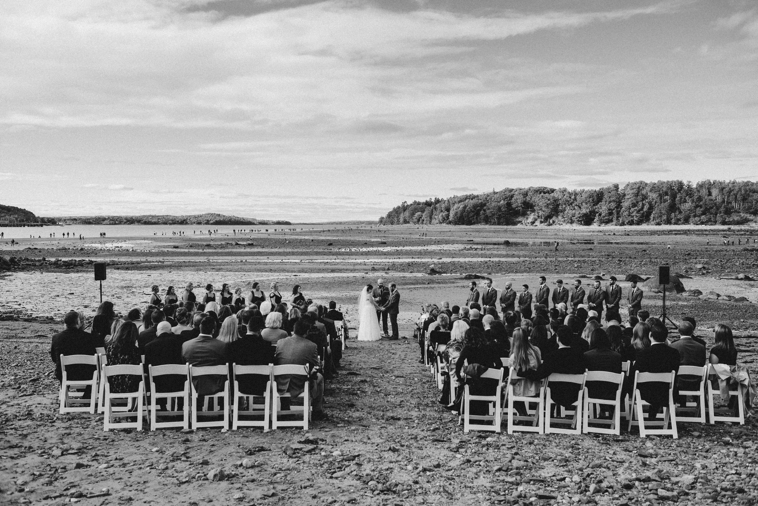 A group of people attending a wedding ceremony on a beach, with the bride and groom standing in front of an officiant, surrounded by bridesmaids and groomsmen, with guests seated in white chairs facing the water and distant trees and people on the sh