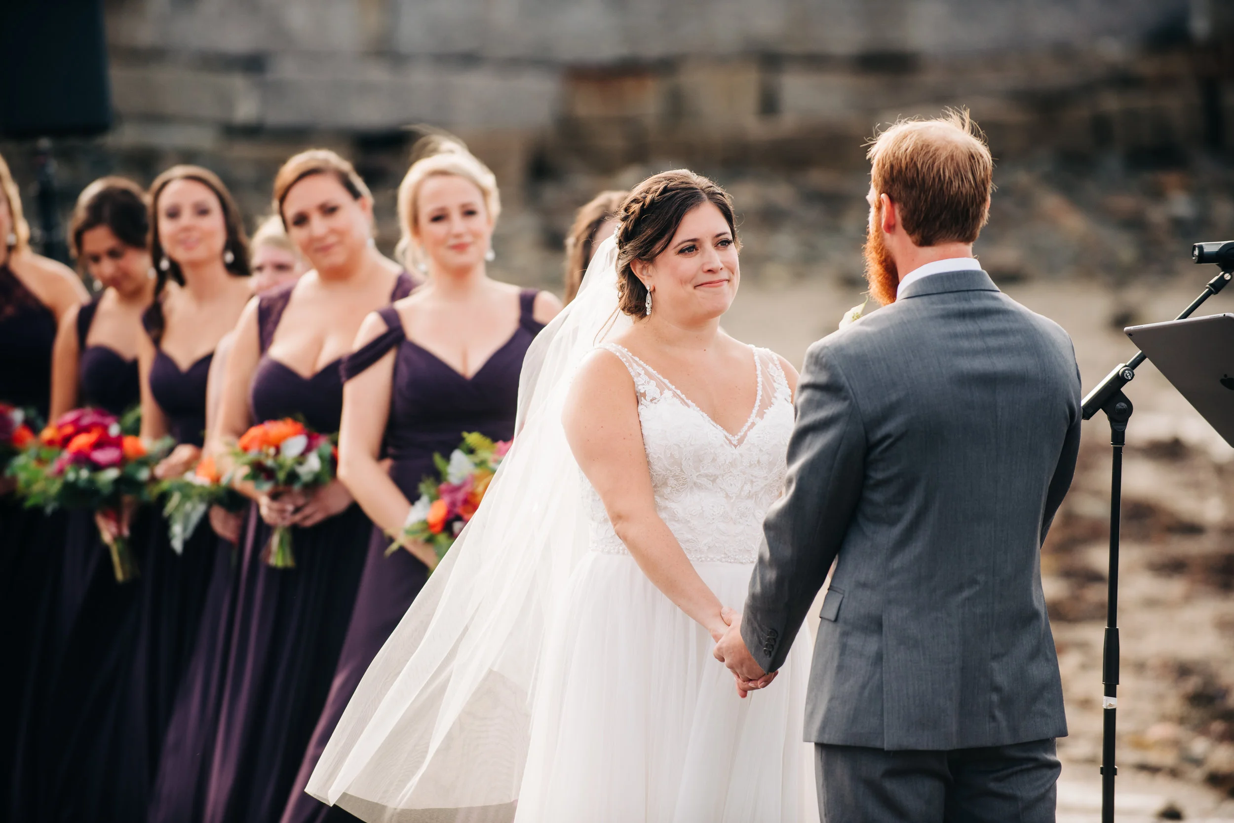 A bride and groom hold hands during their wedding ceremony outdoors, with bridesmaids in purple dresses holding bouquets in the background.