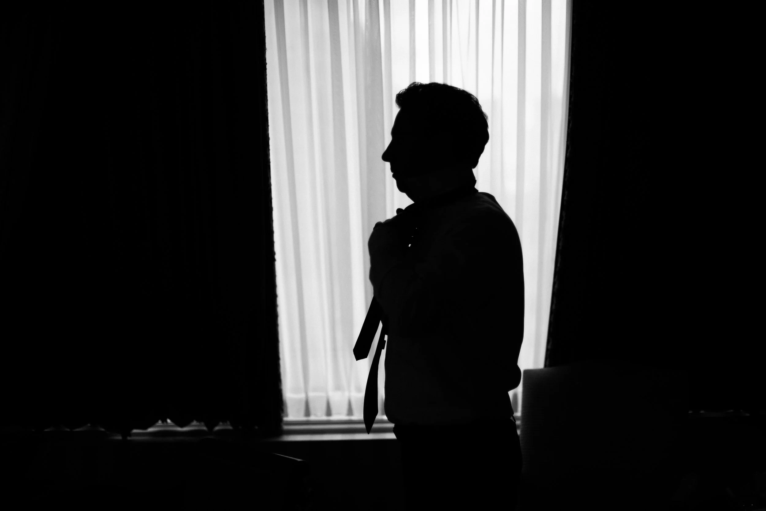 Silhouette of a man with a tie adjusting his collar in front of a window with vertical blinds.
