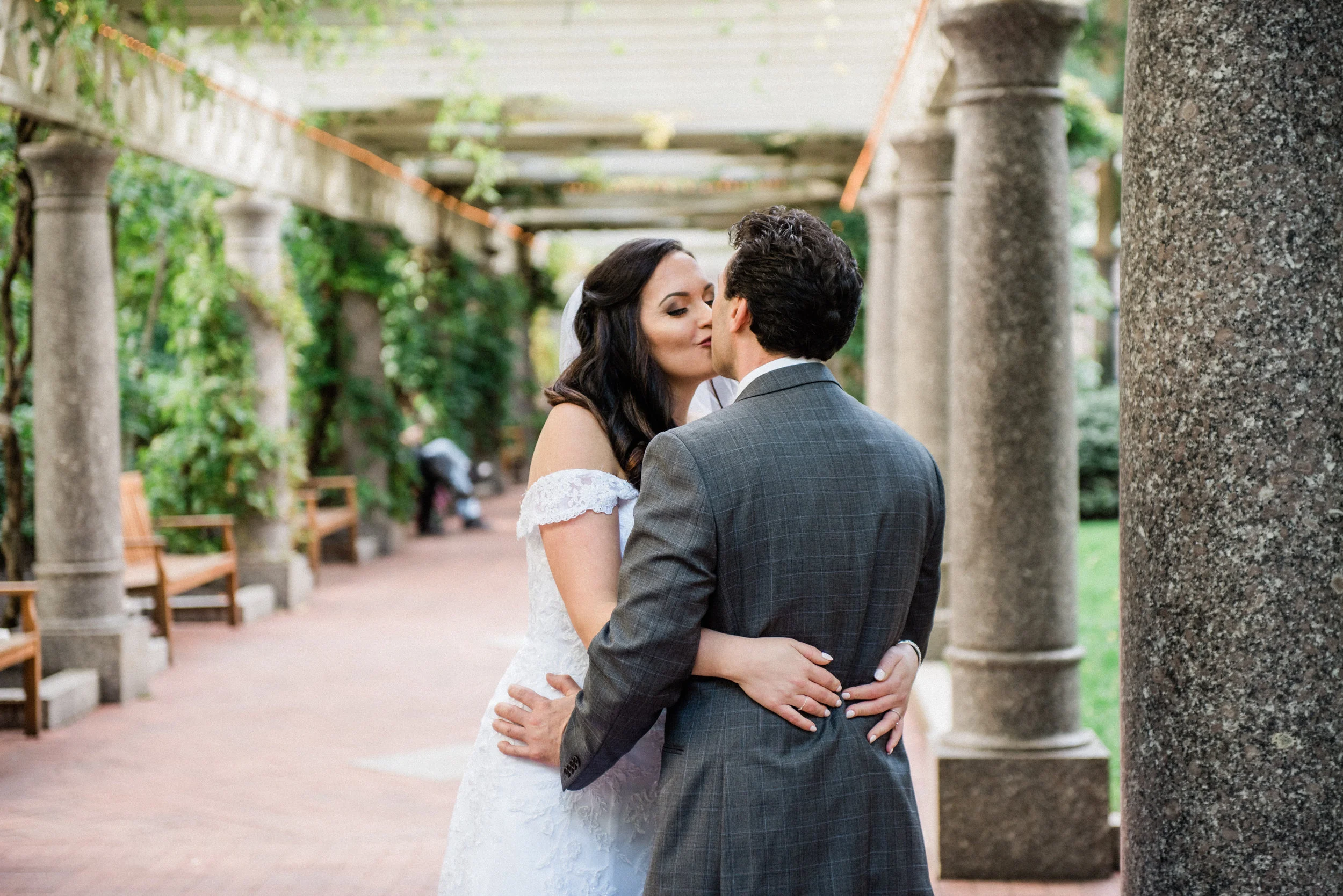 A bride and groom sharing a kiss outdoors surrounded by stone columns and greenery