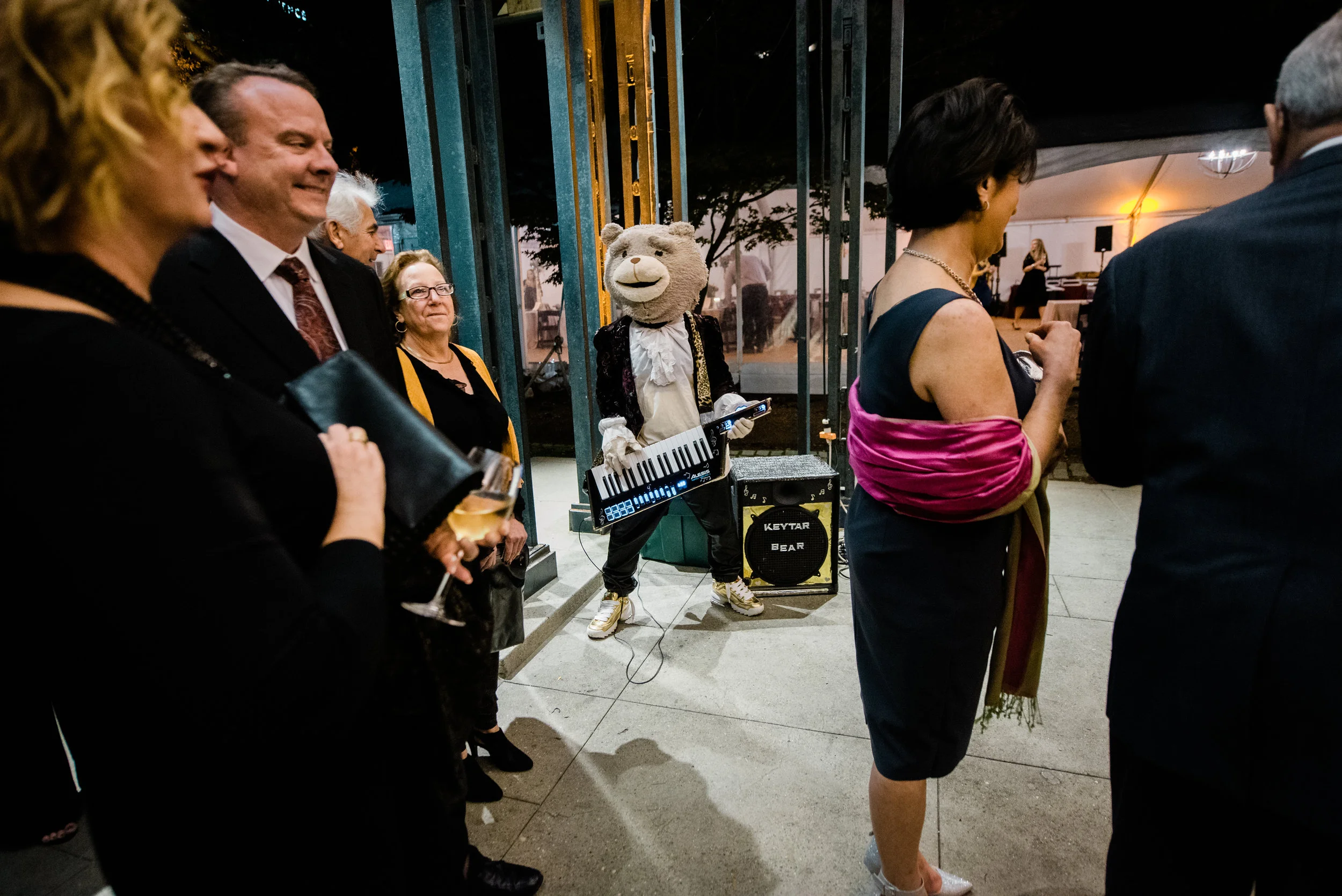 People at a social gathering watching a mascot dressed as a bear playing a keyboard, with some holding drinks, in an indoor-outdoor venue at night.