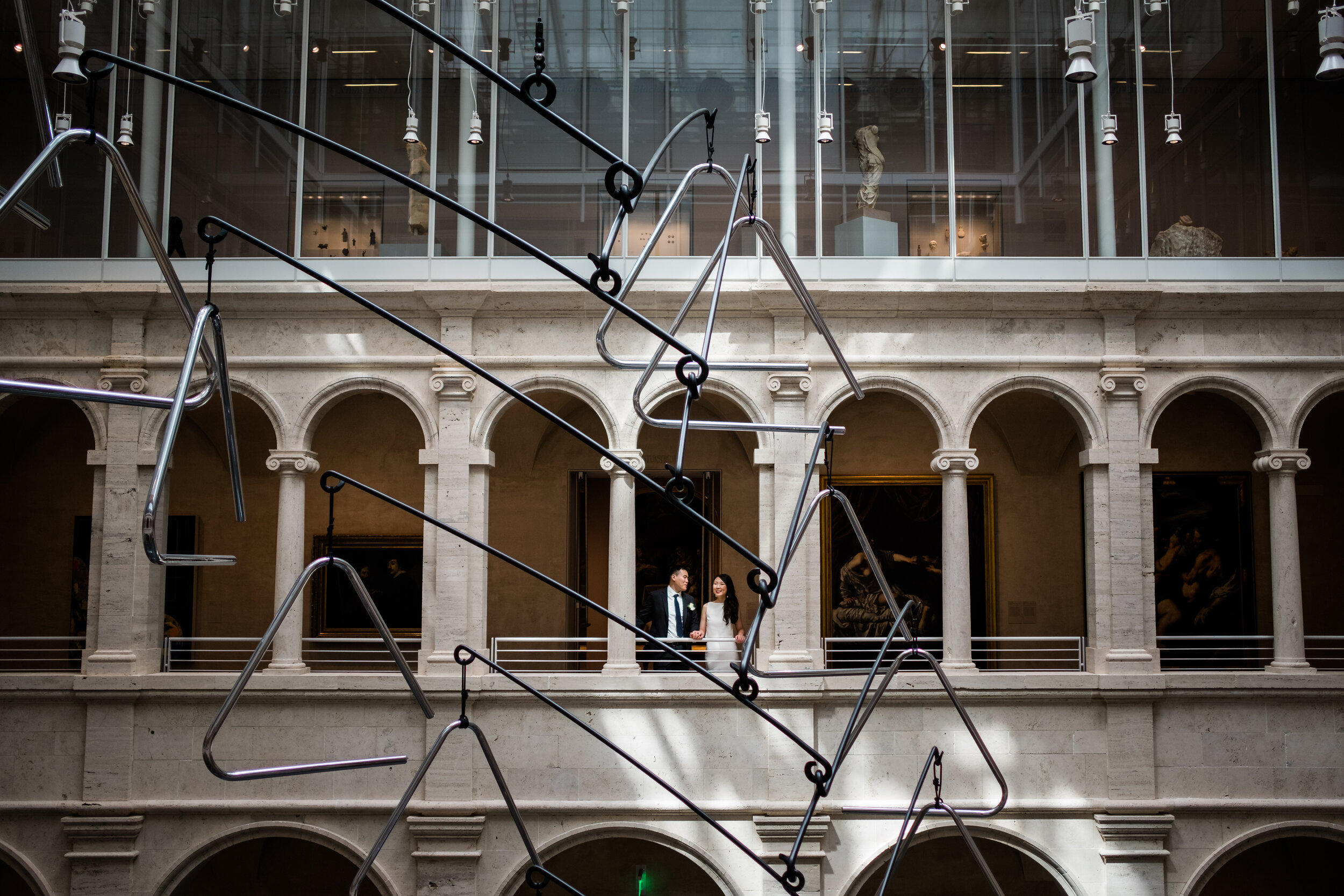 A couple in wedding attire standing on an upper balcony inside a museum, with modern metal sculptures in the foreground and classical architecture with arched windows and framed paintings in the background.