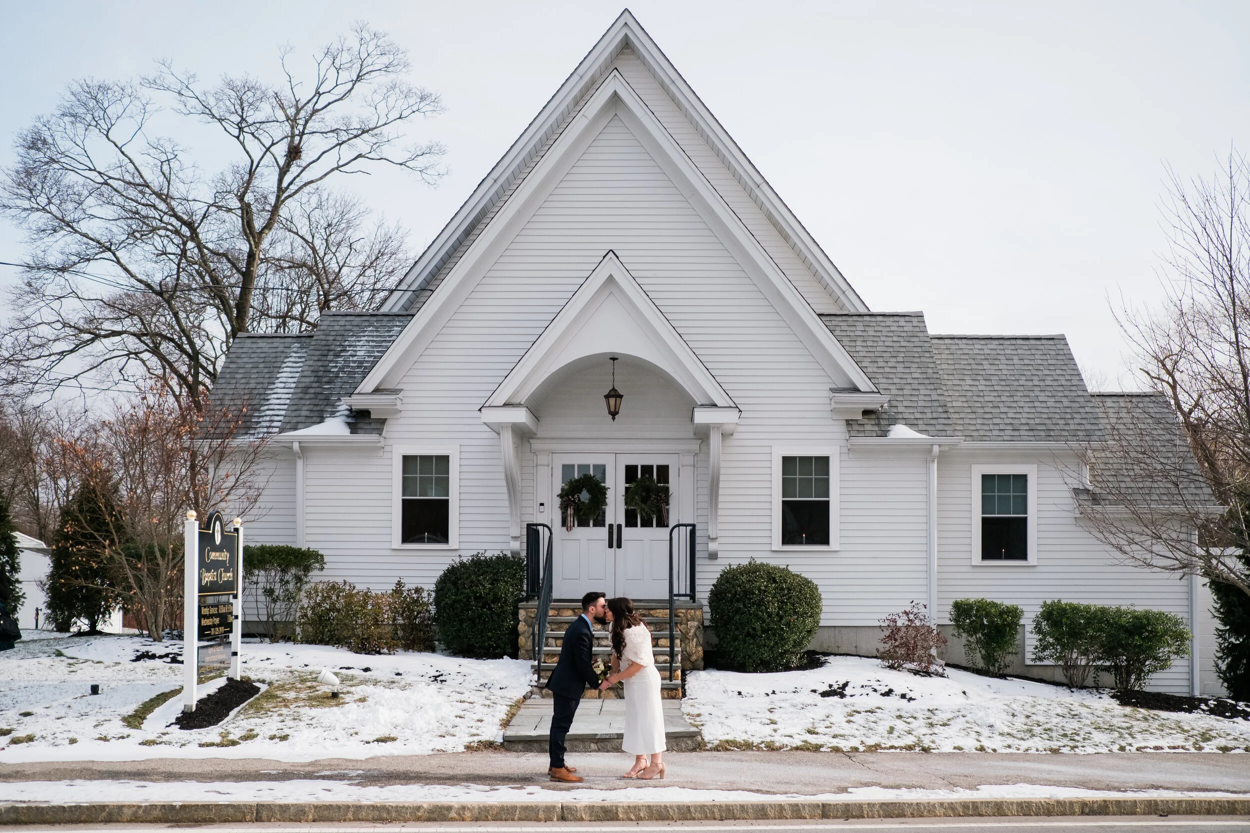 A couple dressed in wedding attire in front of a small white church with snow on the ground, holding hands and sharing a kiss.