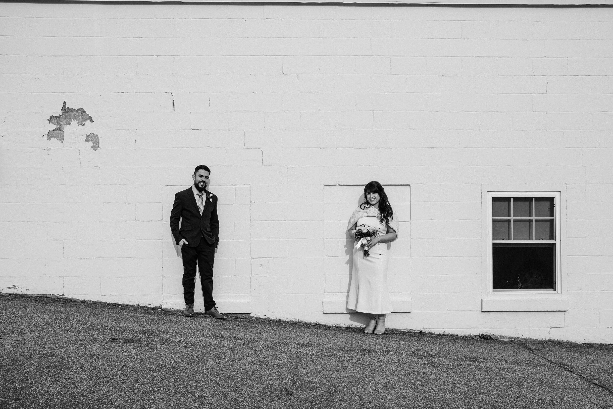 A black-and-white photo of a man in a suit and a woman in a dress standing apart against a large white brick wall, with a small window nearby.