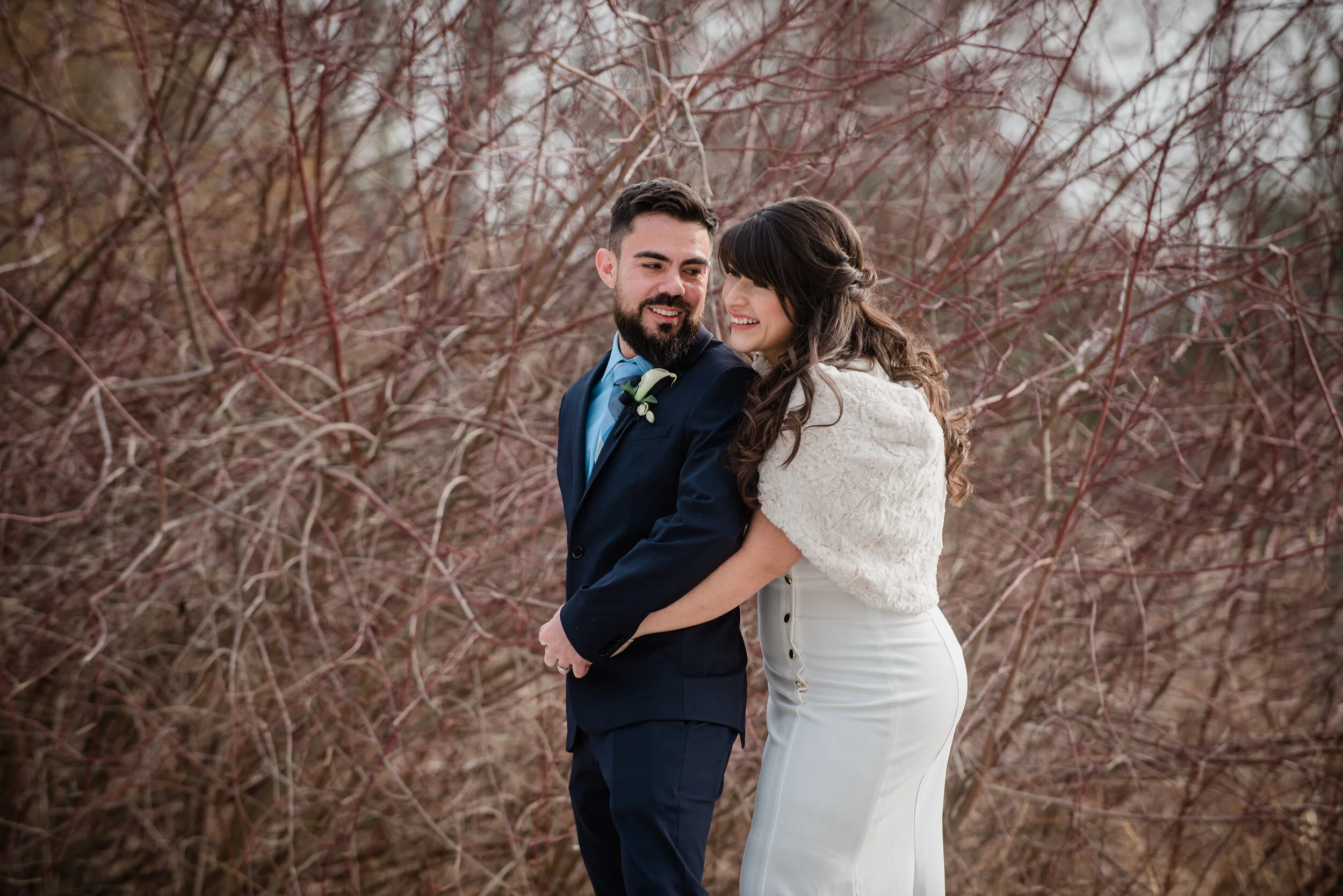 A happy couple on their wedding day standing outdoors in front of leafless bushes, with the bride wearing a white dress and a fluffy white shawl, and the groom wearing a dark suit and a boutonniere.