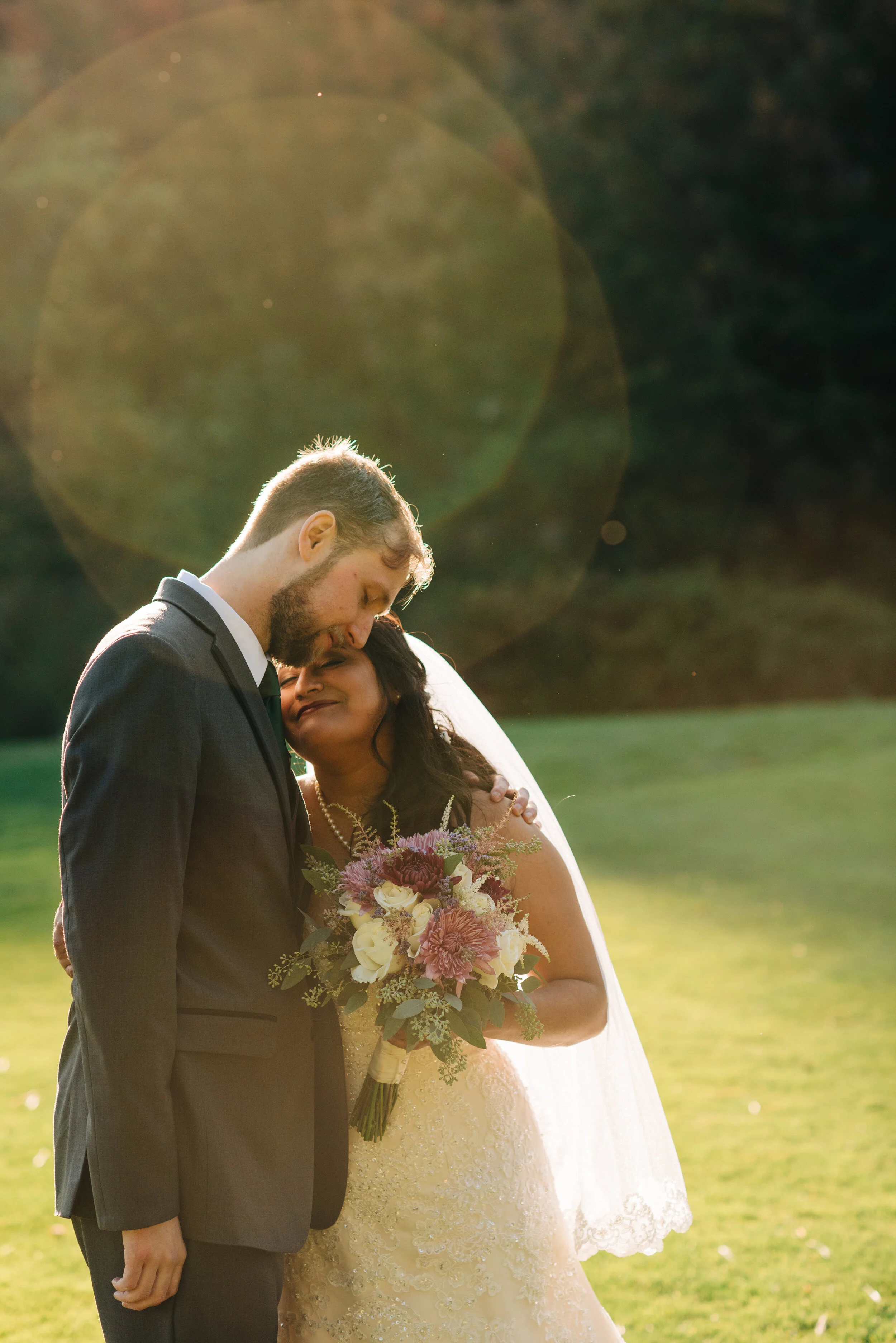 A bride and groom embrace outdoors on their wedding day, with the bride holding a bouquet of flowers and the sun shining behind them, creating a warm glow and lens flare.
