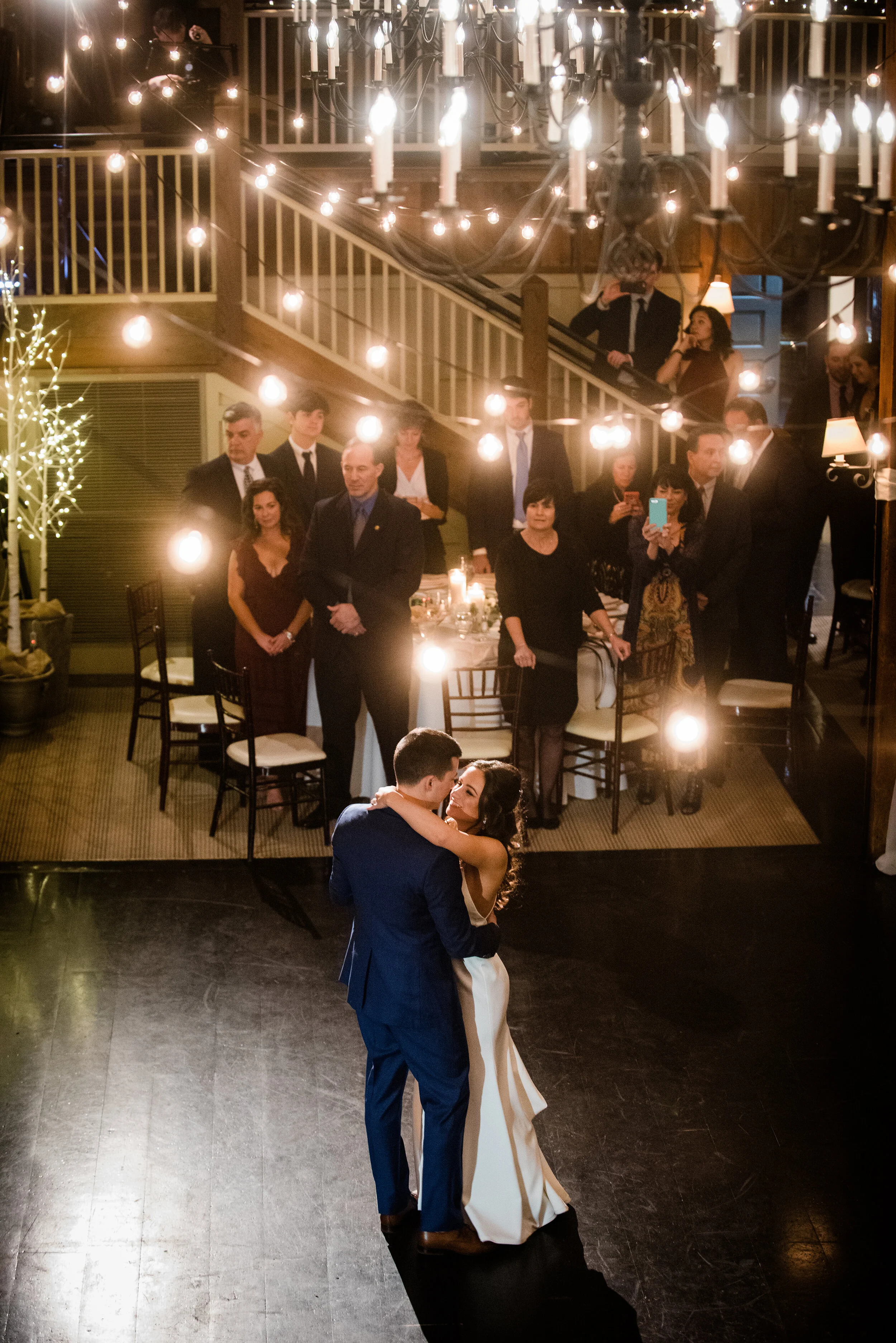 A bride and groom dance closely on a dark dance floor illuminated by soft lighting, while guests watch from a well-lit, elegantly decorated dining area behind them.