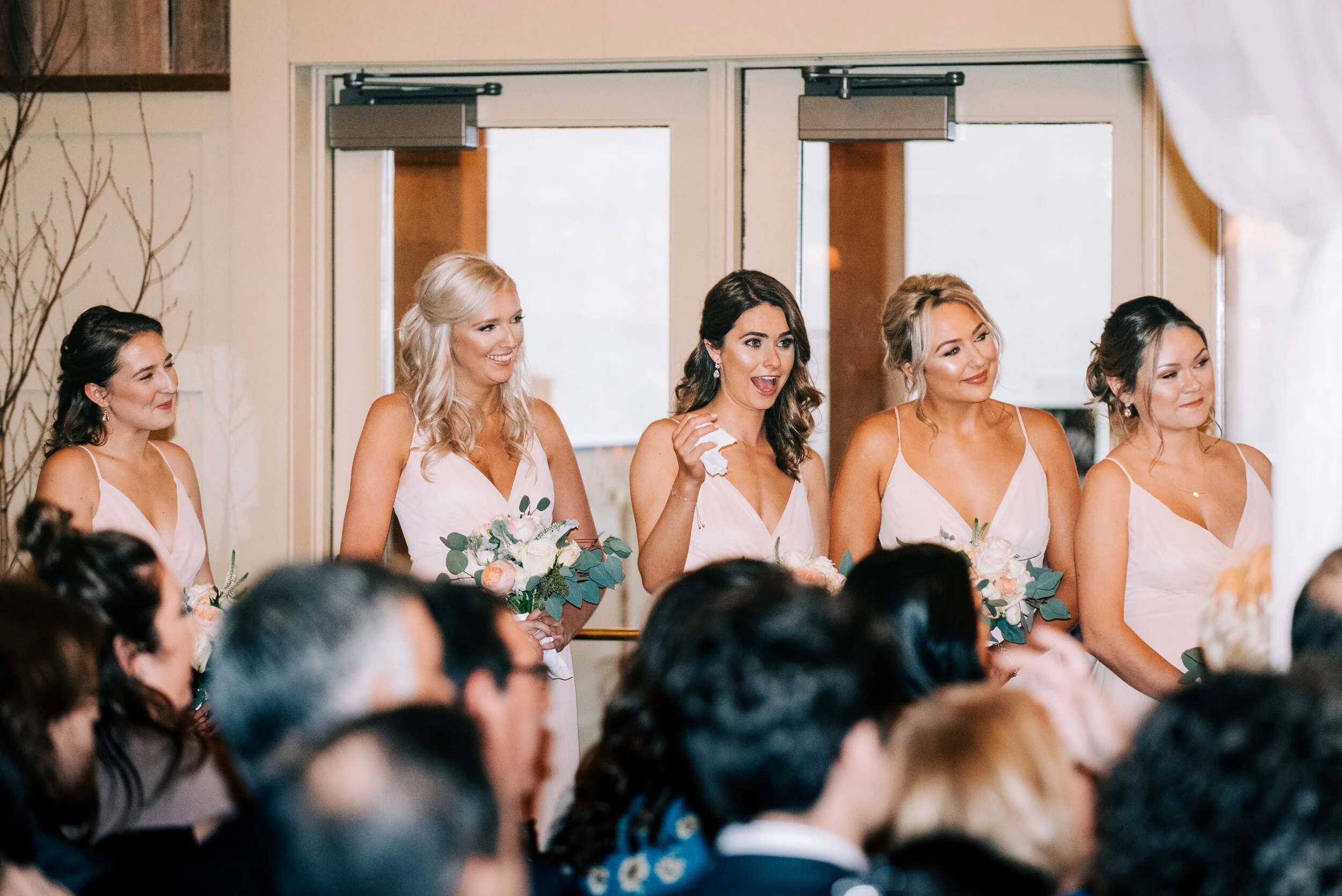 Bride and bridesmaids at a wedding ceremony, standing in a row, holding bouquets of flowers, in front of an audience.