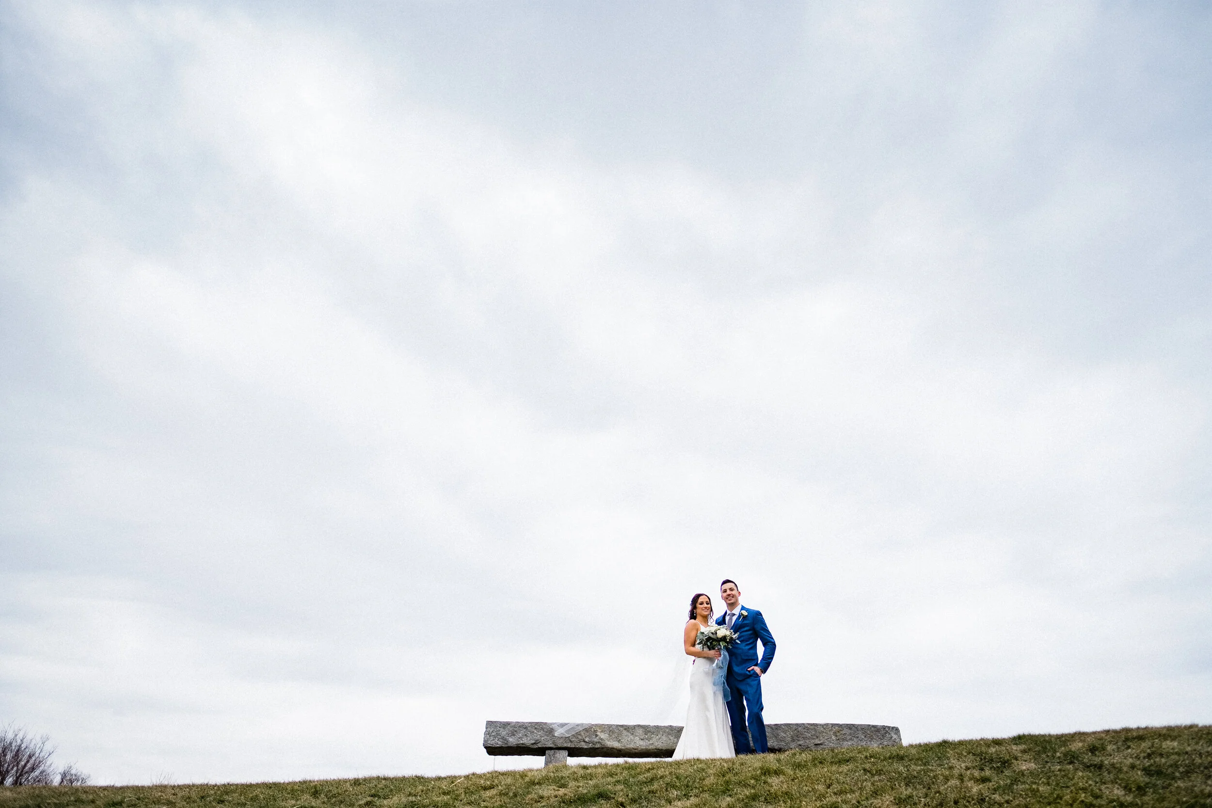 A bride and groom standing together on a grassy hill with a stone bench behind them, under a cloudy sky.