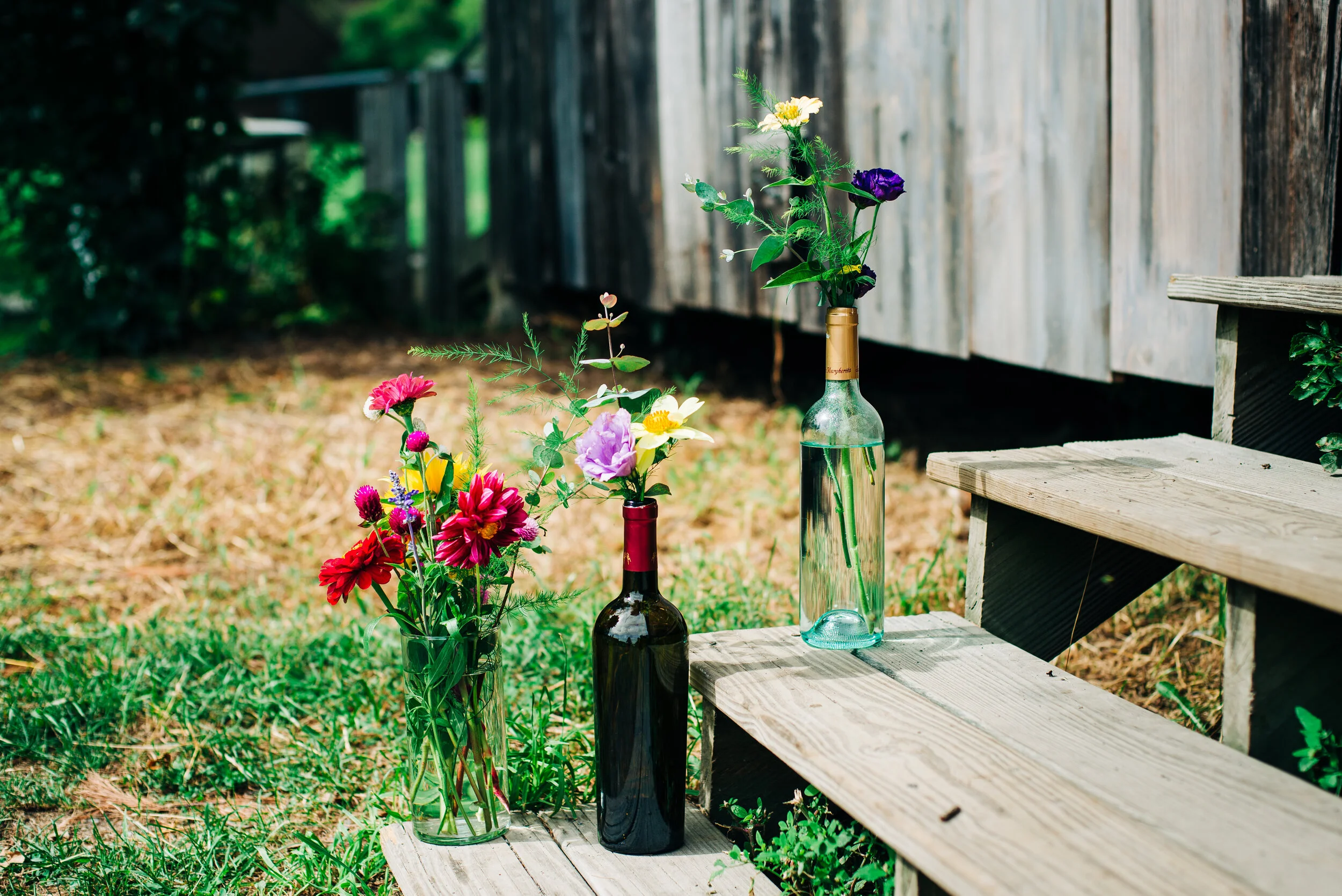 Three glass bottles with colorful flowers placed on wooden stairs outdoors near a weathered wooden shed.