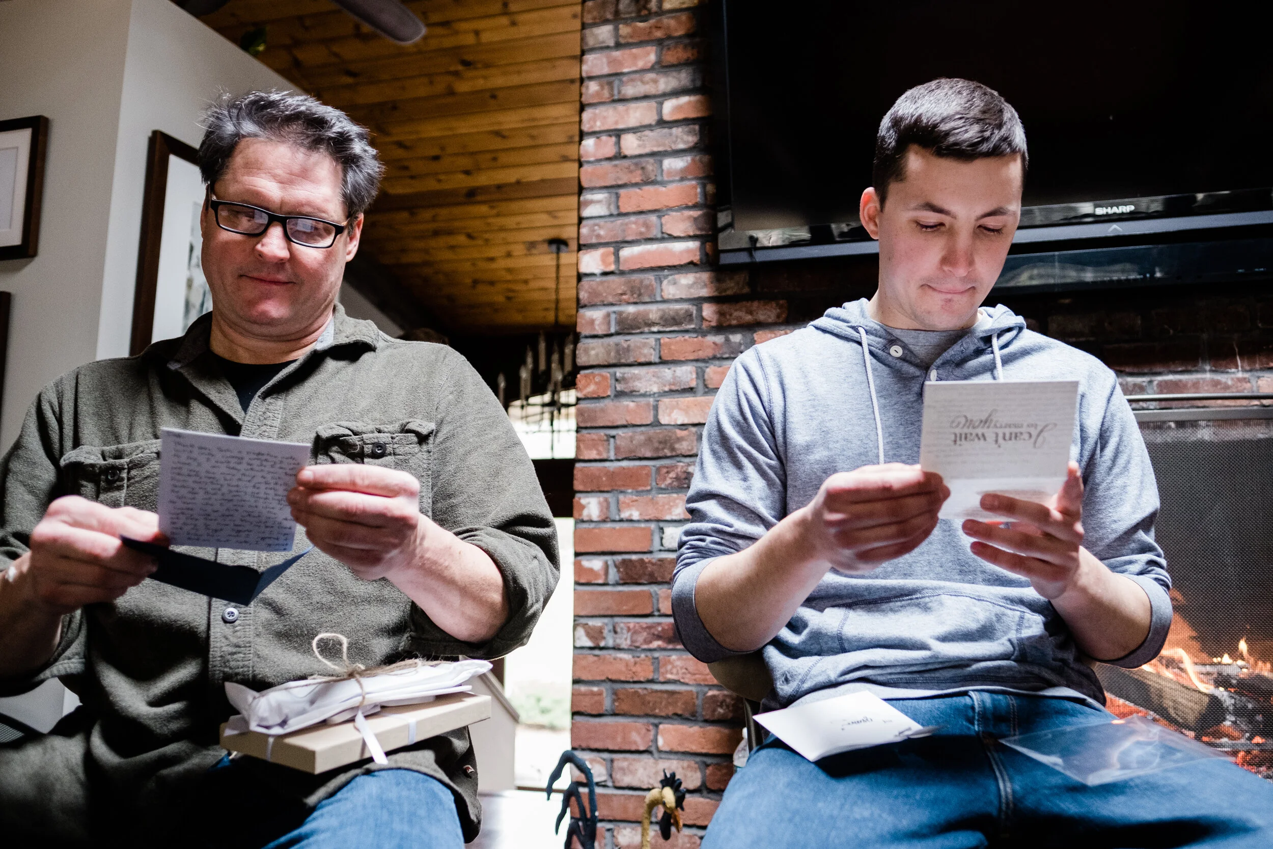 Two men sitting indoors, reading greeting cards. The man on the left wears glasses and a green shirt, holding a card and a wrapped gift. The man on the right wears a gray hoodie, holding a card. A fire burns in a fireplace behind him.