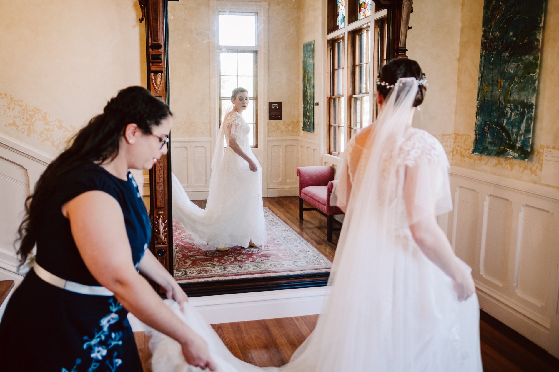 A woman in a wedding dress looking into a large ornate mirror, with her reflection visible, while another woman helps her with her dress in a warmly lit room with wooden floors and art on the walls.